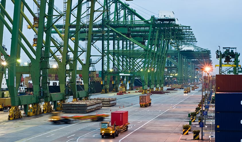 Cranes and trucks operating at a brightly lit container port in the evening.