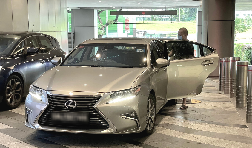 A person wearing a mask enters a silver Lexus private-hire car parked at a sheltered pickup point.