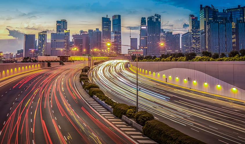Long-exposure view of a busy expressway with light trails from vehicles leading into a modern city skyline at dusk.