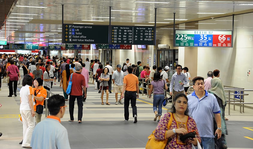 Crowded indoor bus interchange with commuters walking toward various boarding berths.