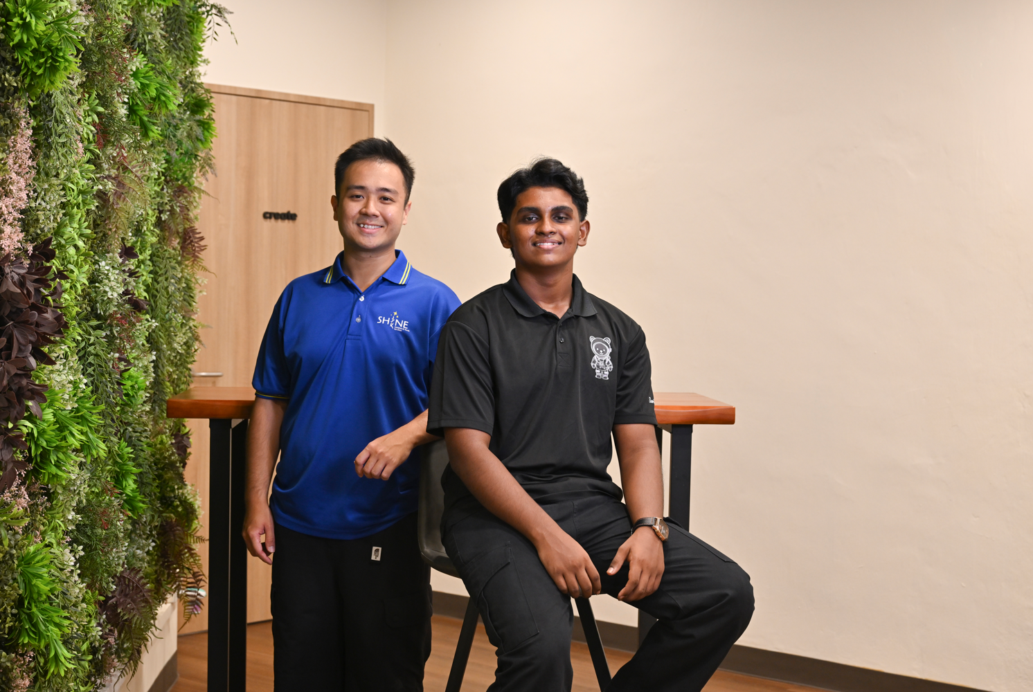 Two young men smile for a photo — one standing in a blue SHINE polo, the other seated in a black polo — beside a lush indoor green wall