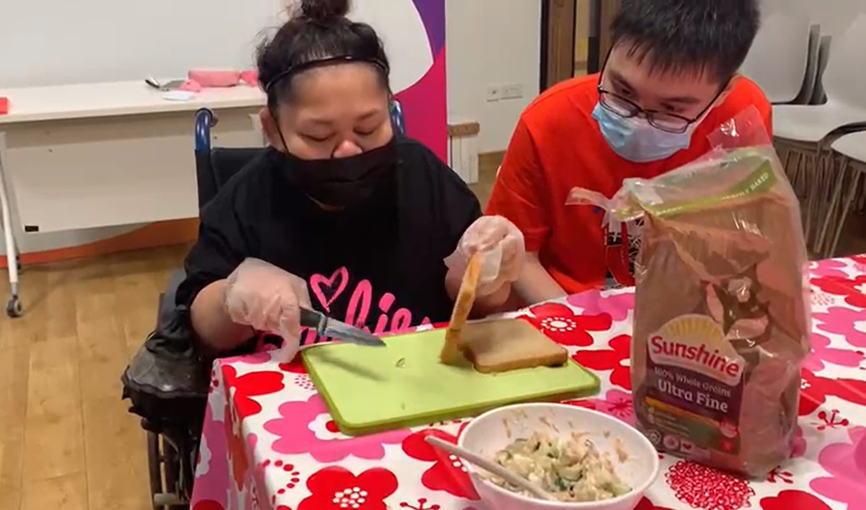 Two people in masks make sandwiches, Sunshine bread on table, salad, cutting board.