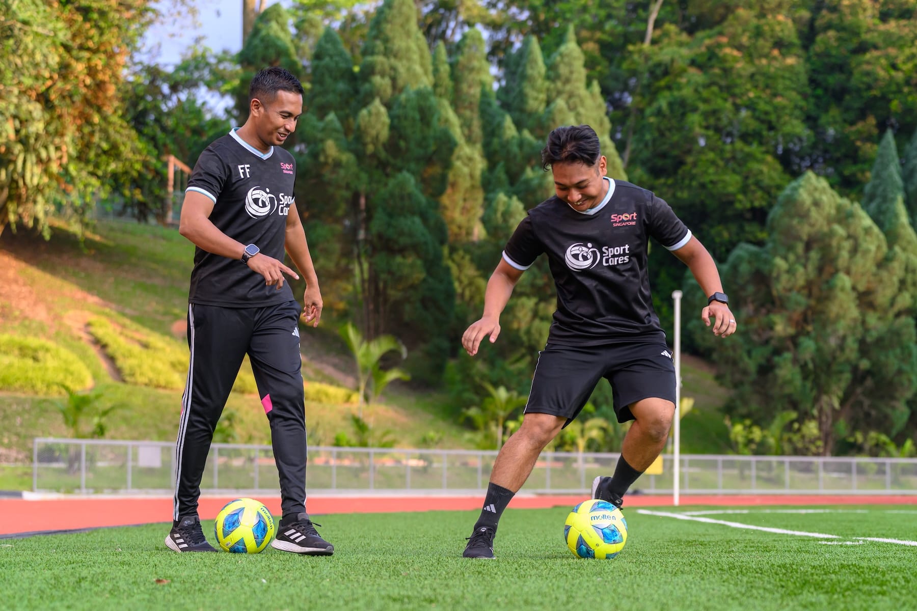 Two men in SportCares shirts practising football on a field, smiling during a training session.