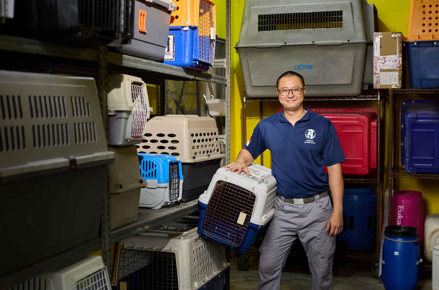 A man in a polo shirt holds an animal carrier, surrounded by shelves stacked with various pet crates