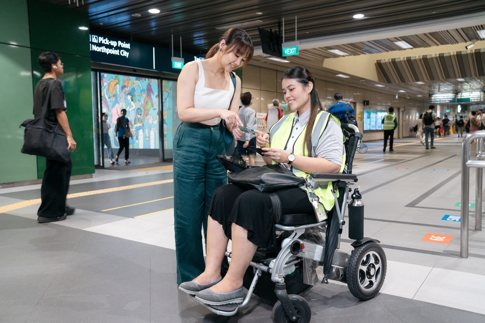 Train station: Woman in wheelchair, wearing safety vest, receiving transit card from another woman.