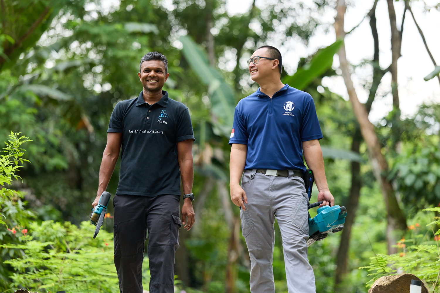 Two men walk together through a lush green outdoor setting, both smiling and carrying handheld equipment. One wears a black ACRES polo shirt, the other a blue polo shirt