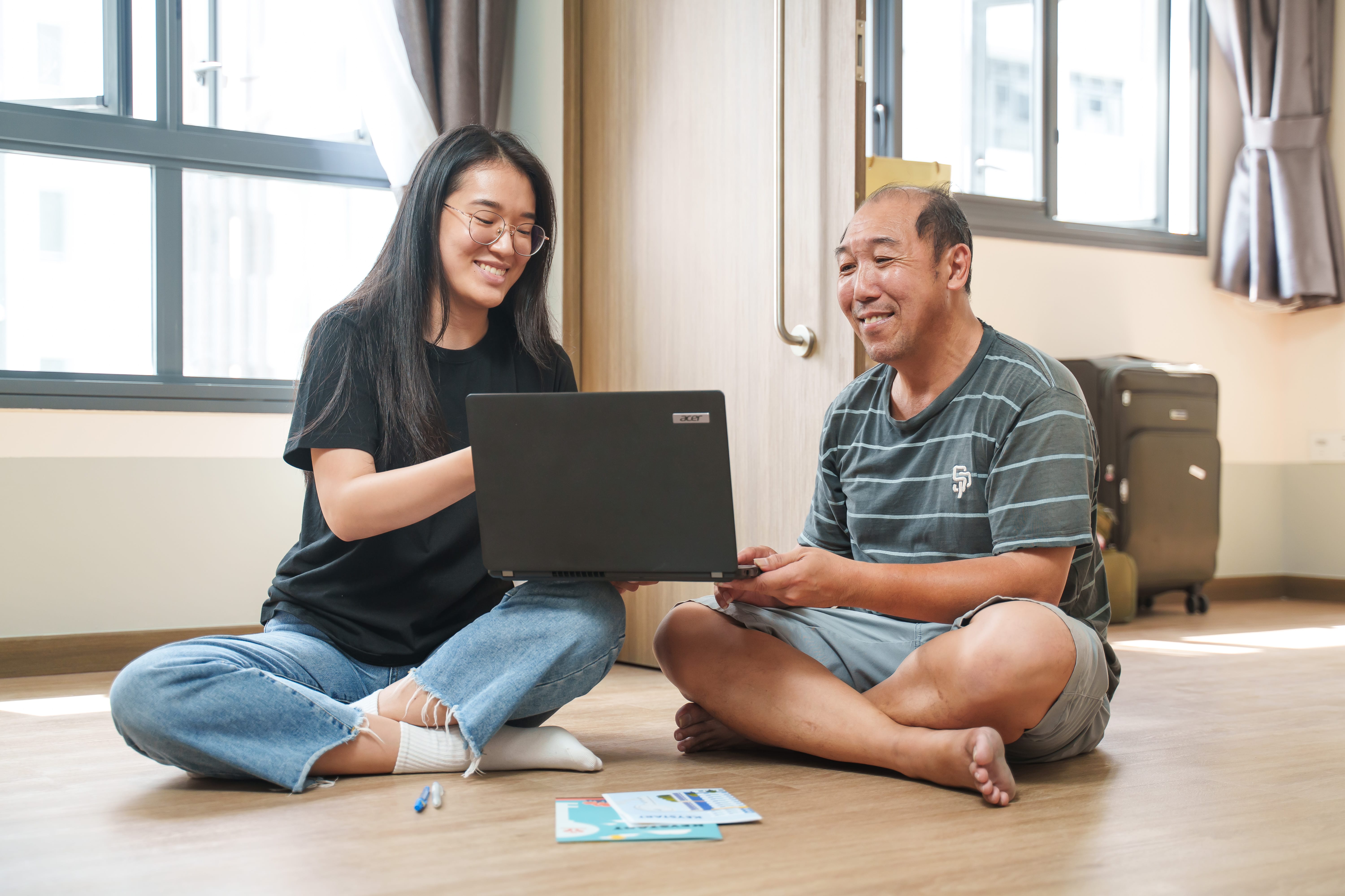 A young lady and a middle-aged man sitting on the floor together, sharing a laptop, with documents placed on the floor.