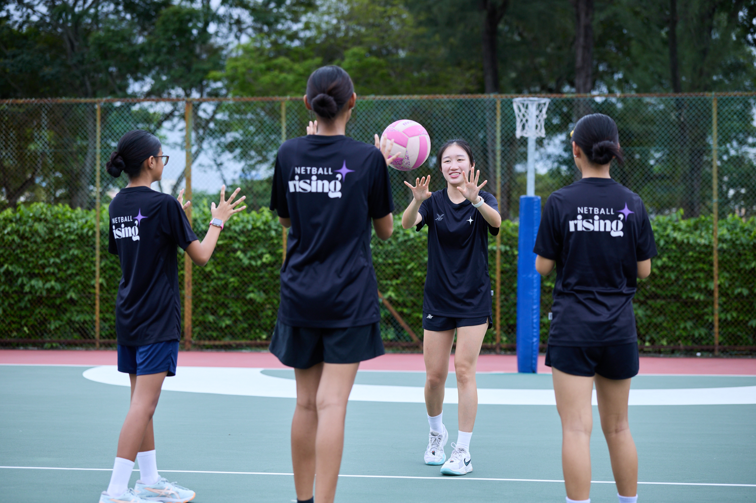 Young players in 'Netball Rising' jerseys practise passing drills on an outdoor court, smiling as the ball flies between them