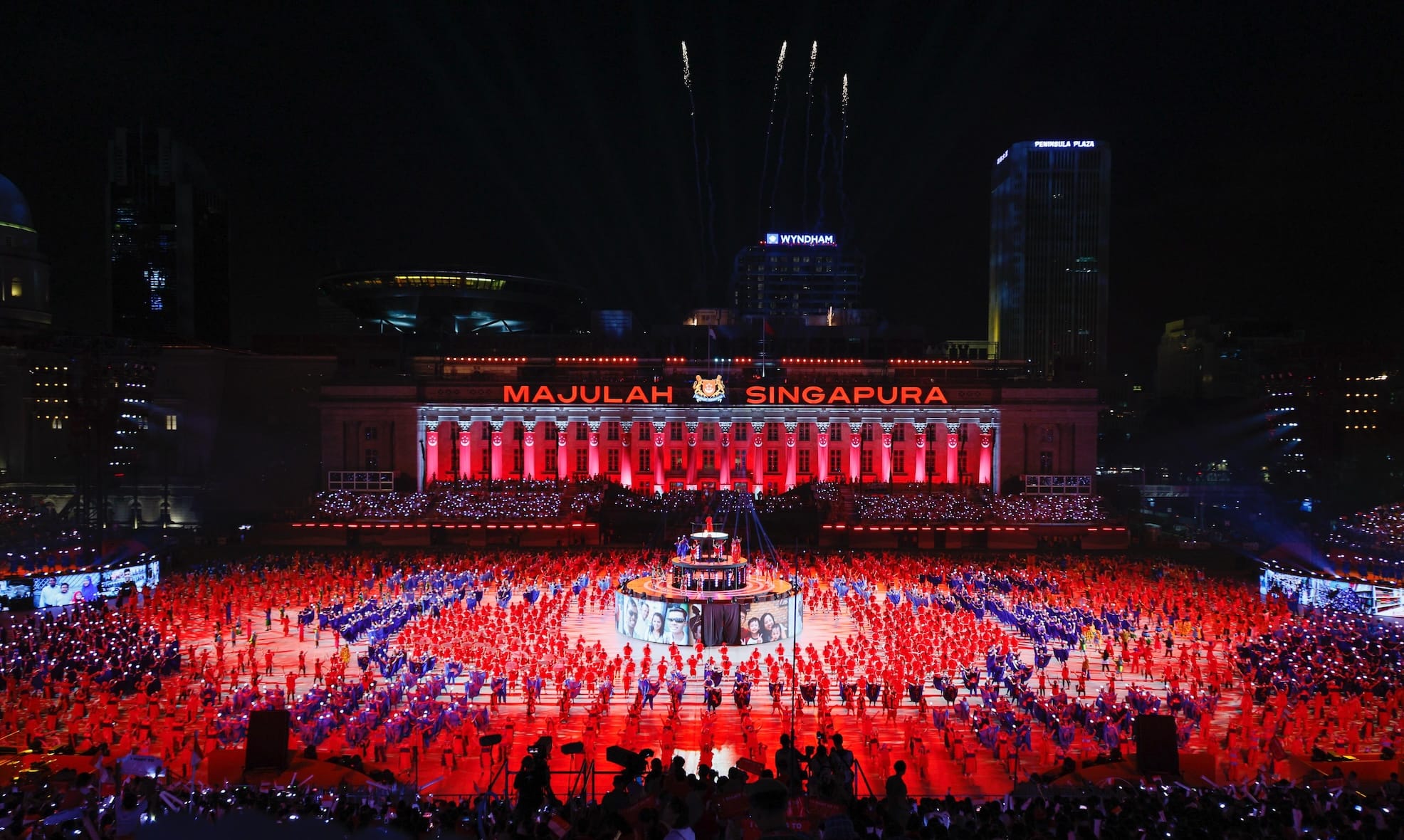 Night view of "Majulah Singapura" building with dancers, fireworks, and hotel signs.