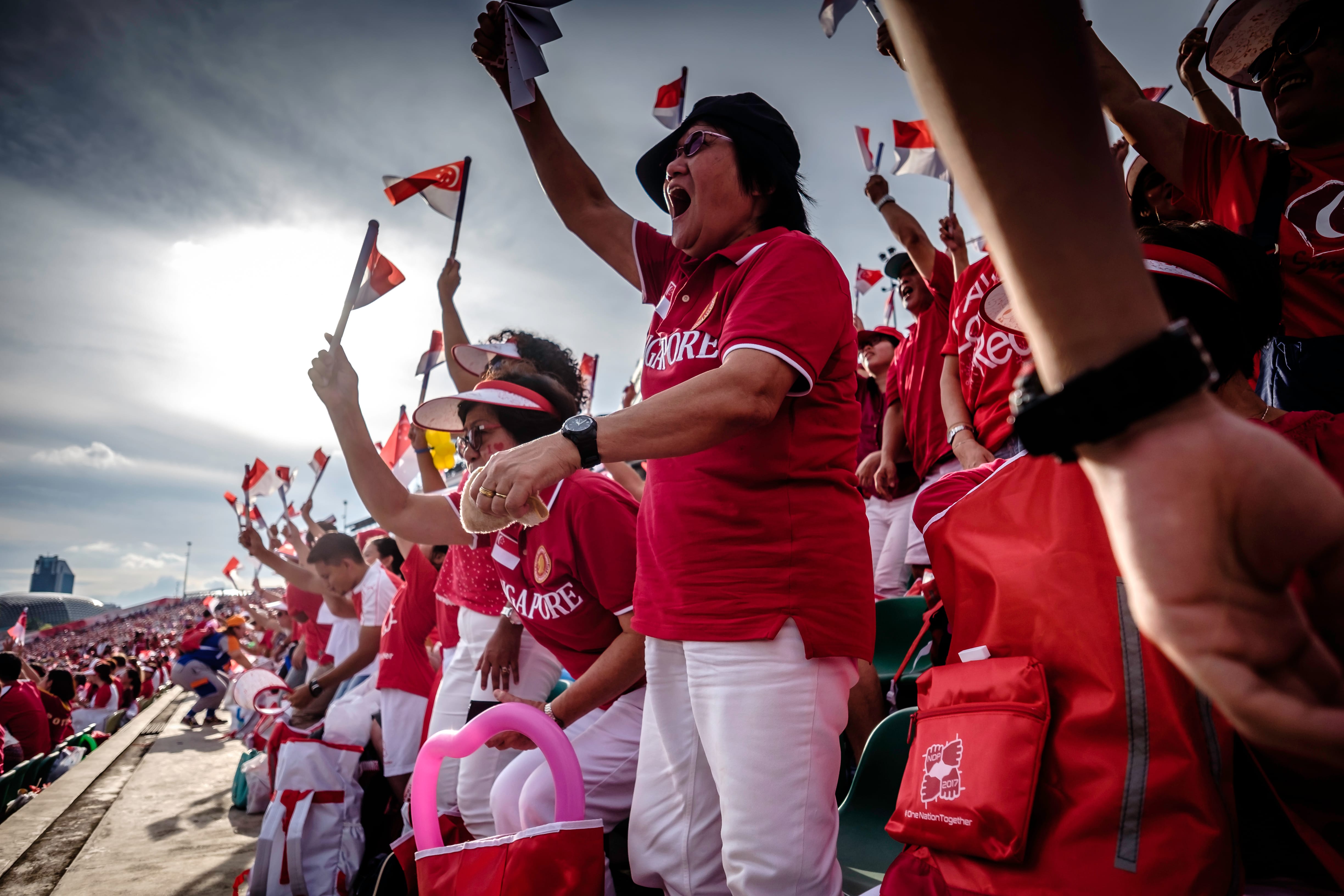 Crowd cheering, waving Singapore flags, wearing red shirts with "Singapore" text.
