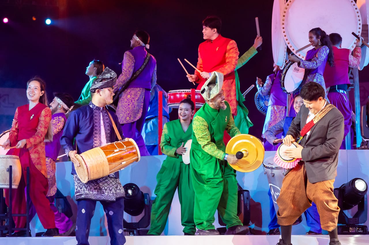 A lively multicultural percussion performance on stage, with musicians in colourful traditional Southeast Asian costumes playing various traditional instruments