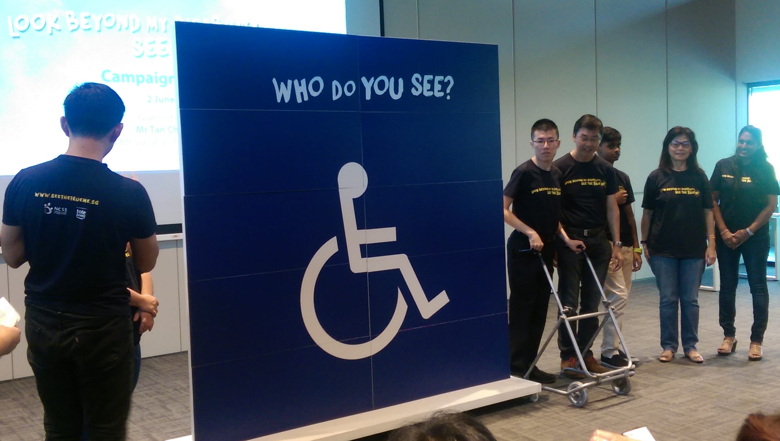 Participants stand beside a large panel showing a wheelchair symbol with the text “Who do you see?” during a disability awareness event.