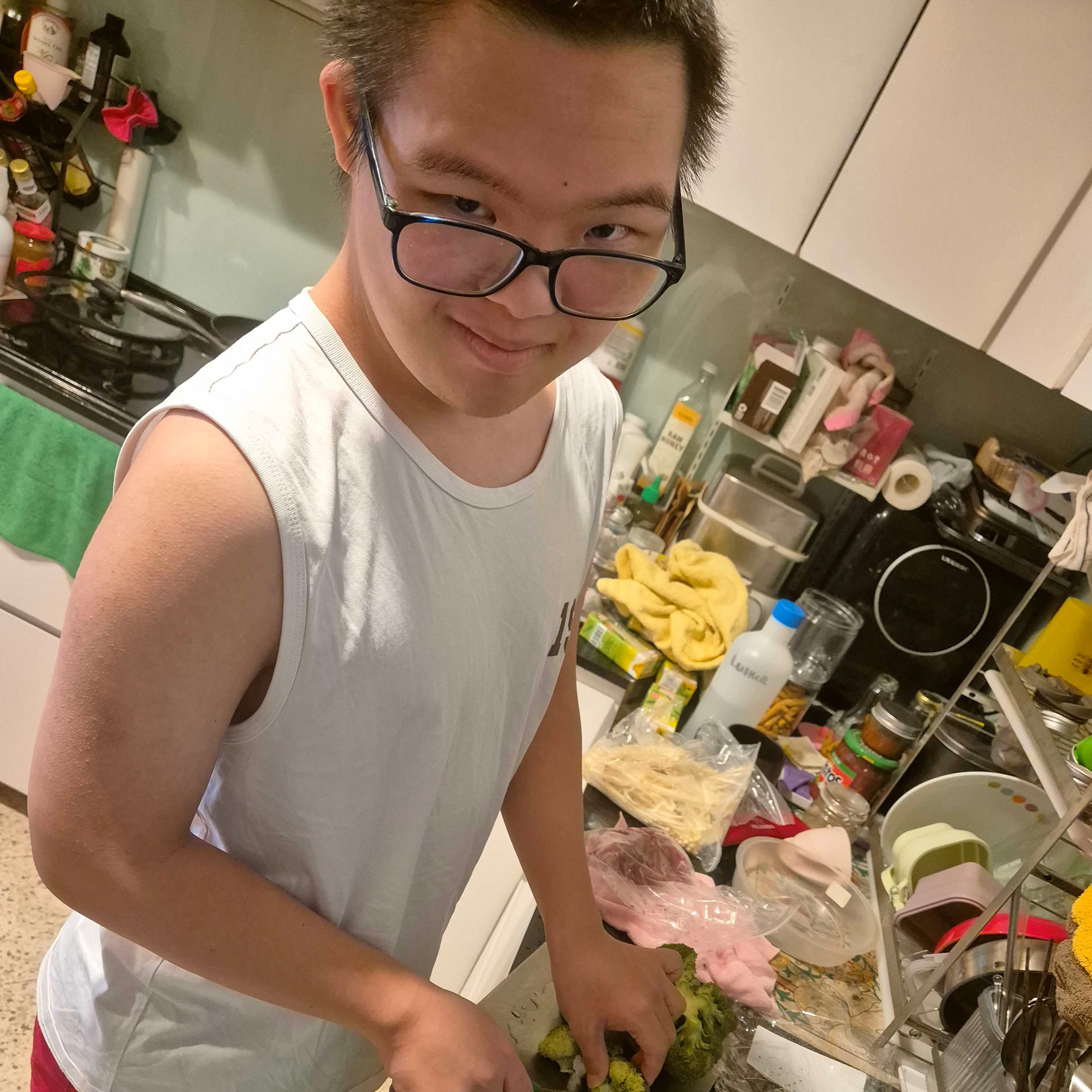 Person in tank top and glasses cuts broccoli on cluttered counter.