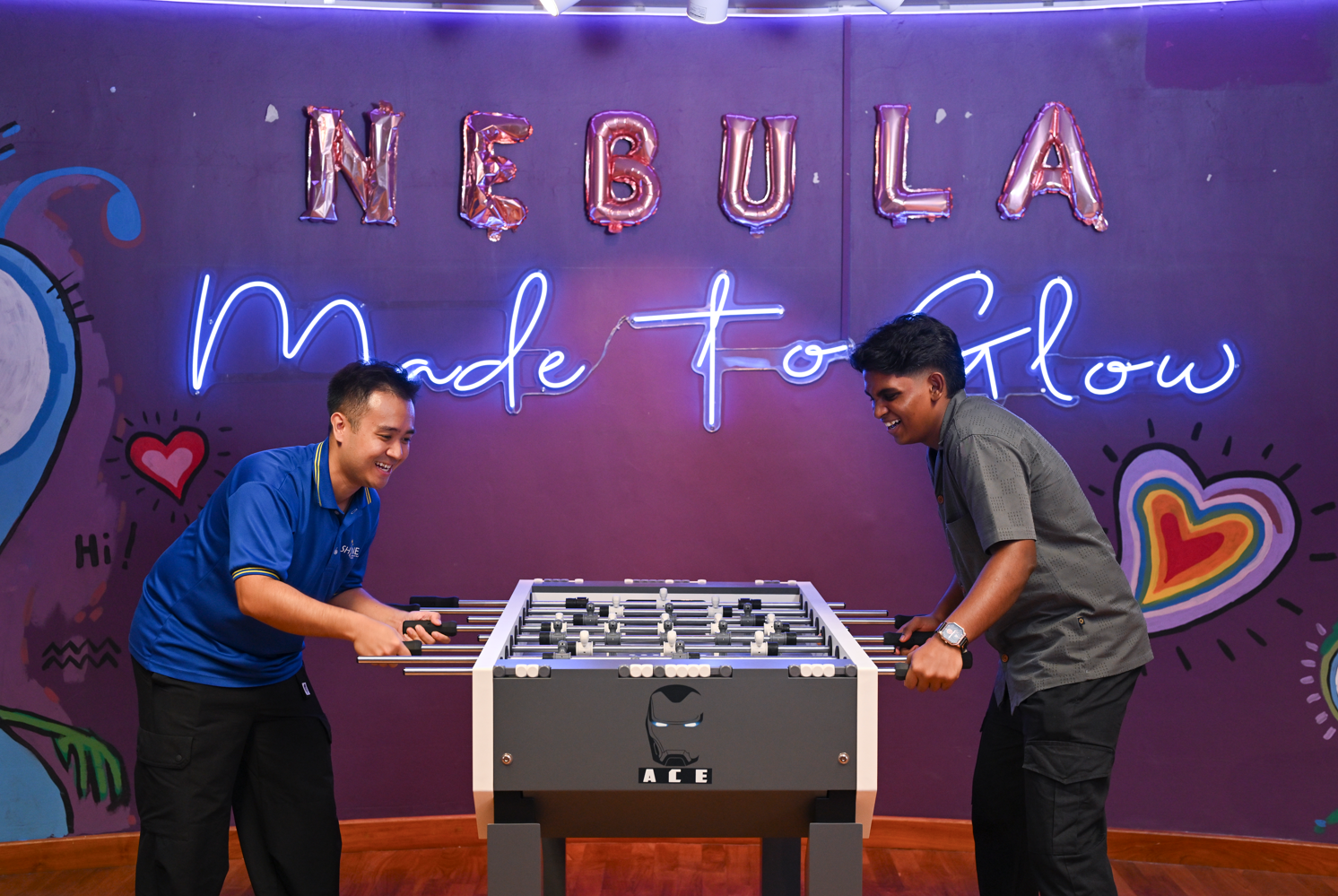 Two young men laugh as they face off over a foosball table, set against a vibrant purple wall with "NEBULA" in balloon letters and a neon "Made to Glow" sign behind them