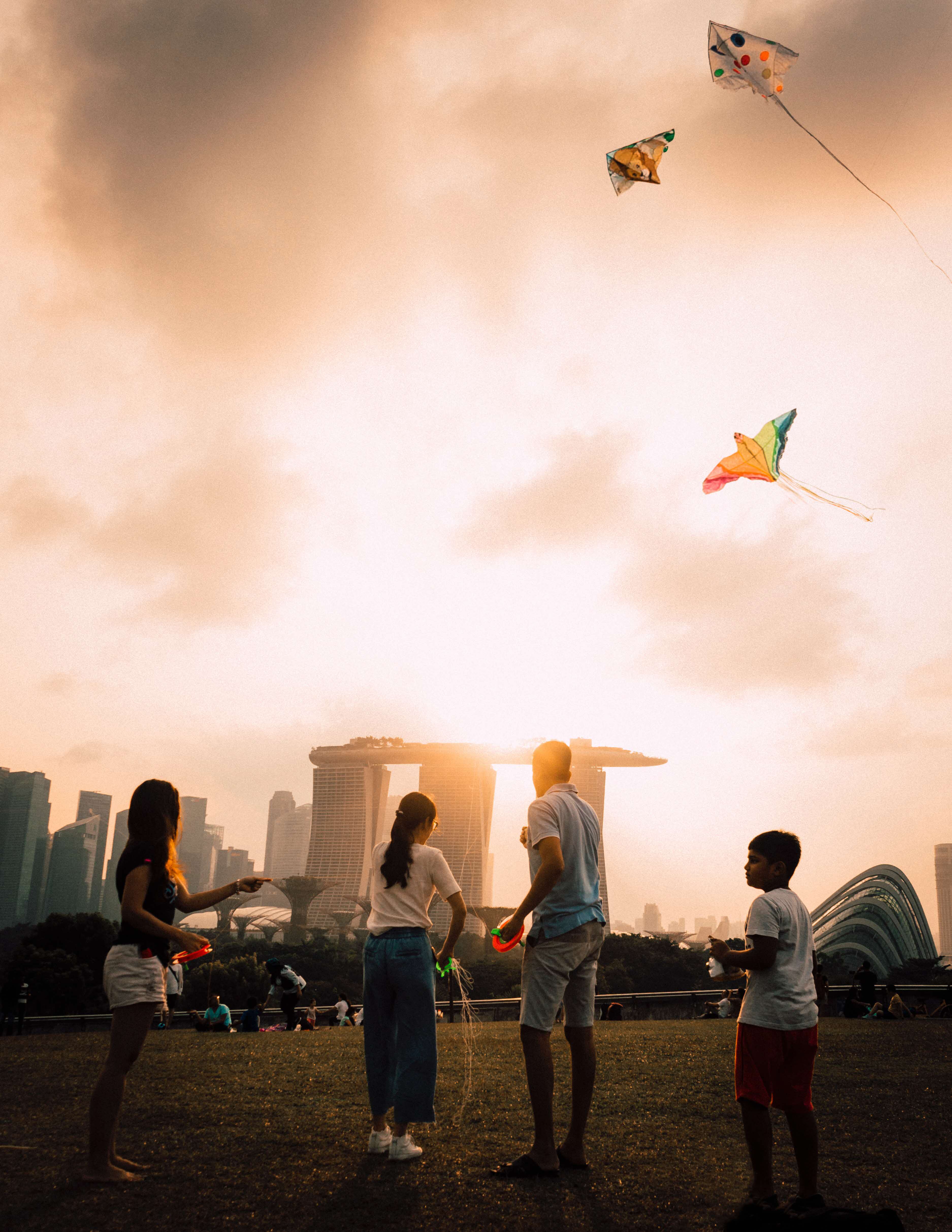 Four people flying kites on grass, with the Singapore skyline and sunset in the background.