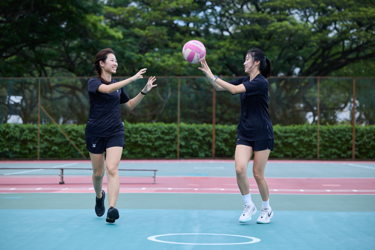 Two players in matching dark uniforms share a smile as they pass a pink and white netball to each other on an outdoor court