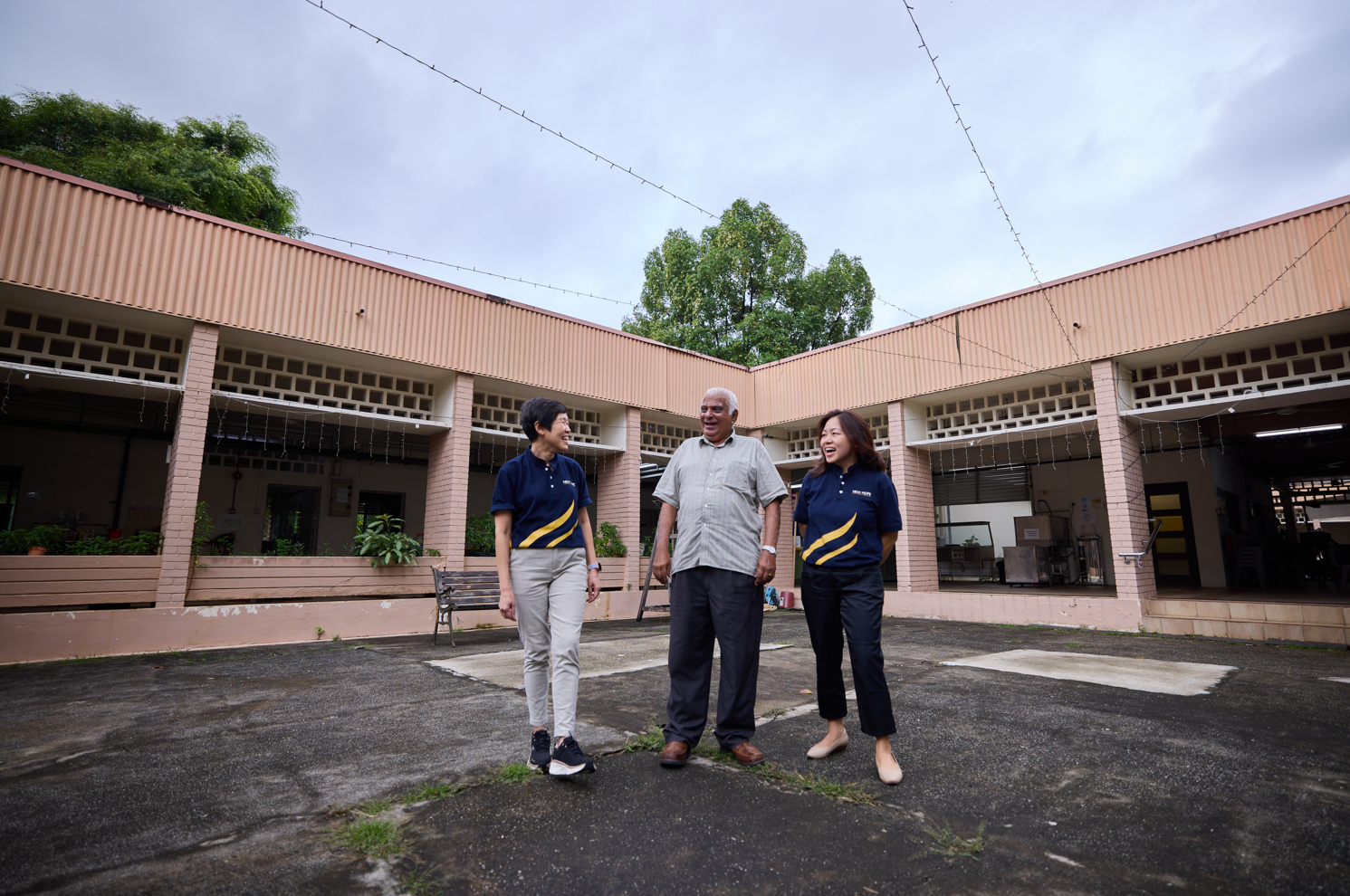 Three people chat warmly while strolling through an open courtyard, two women in matching navy and light orange polos flanking an elderly gentleman