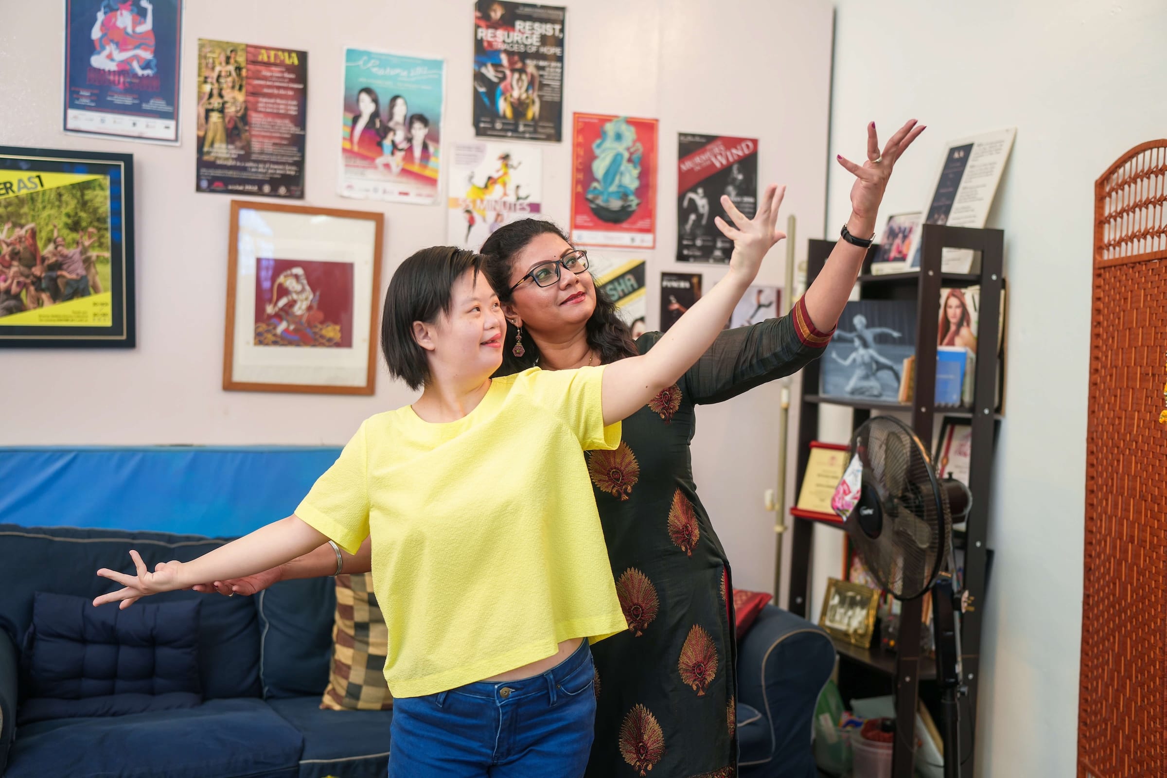 Two women practice dance poses in a room with posters and a blue sofa.