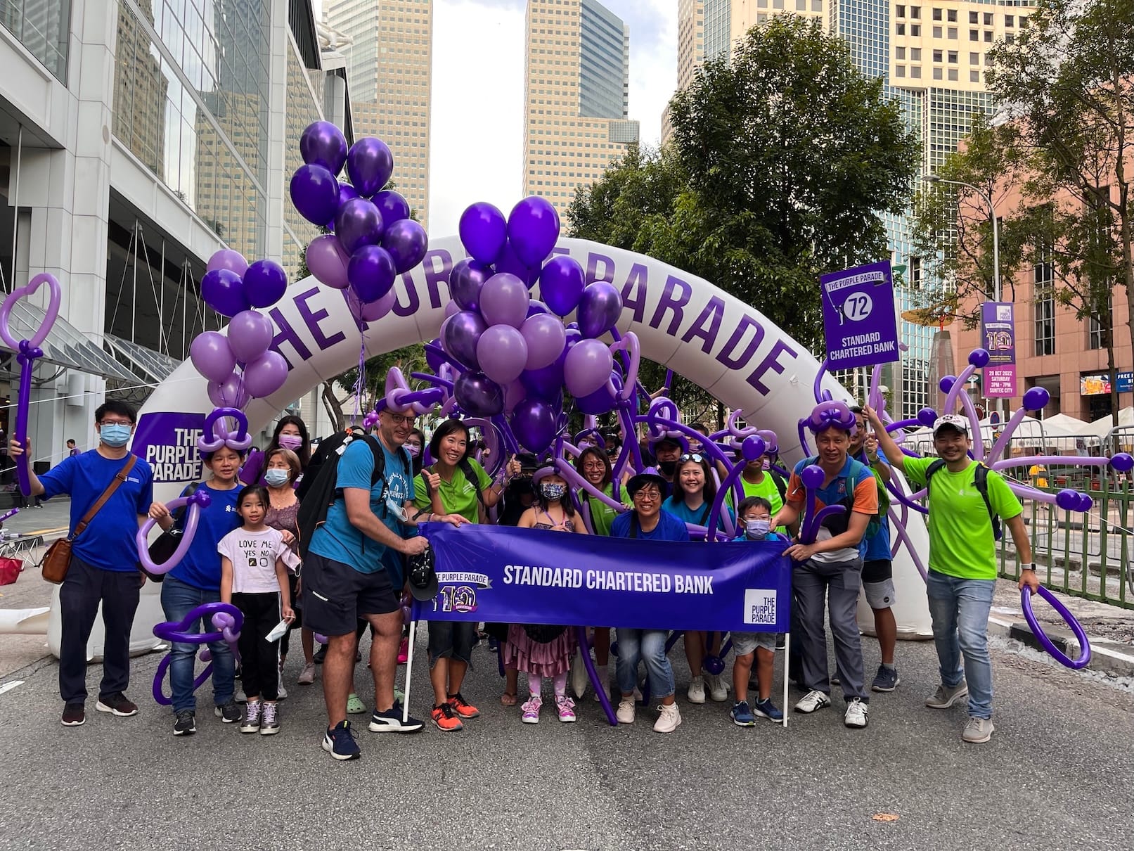 Group at "The Purple Parade" with Standard Chartered banner, balloons, and city buildings.