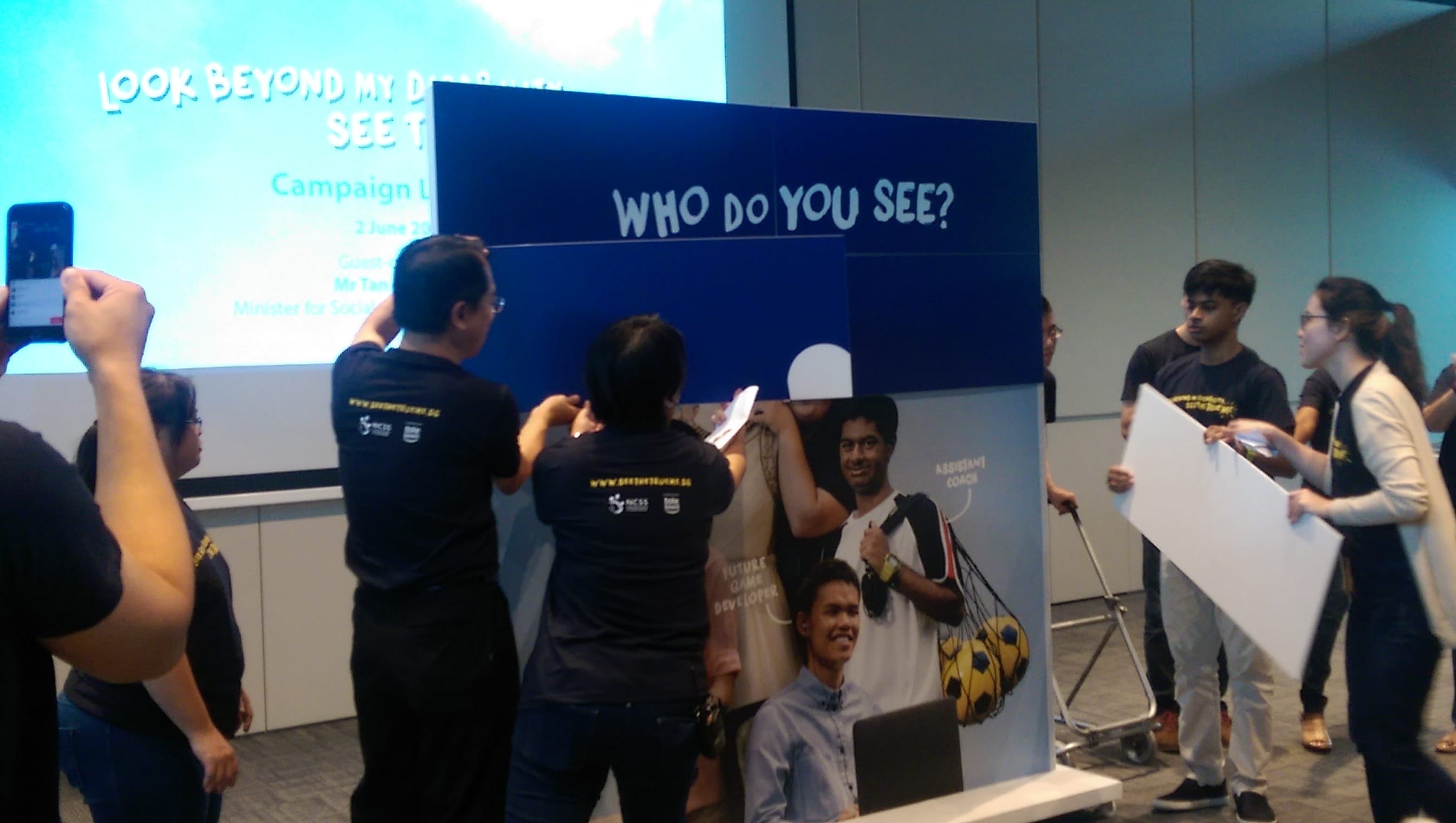 Participants remove panels to reveal portraits behind a display reading “Who do you see?” as part of a disability inclusion campaign.
