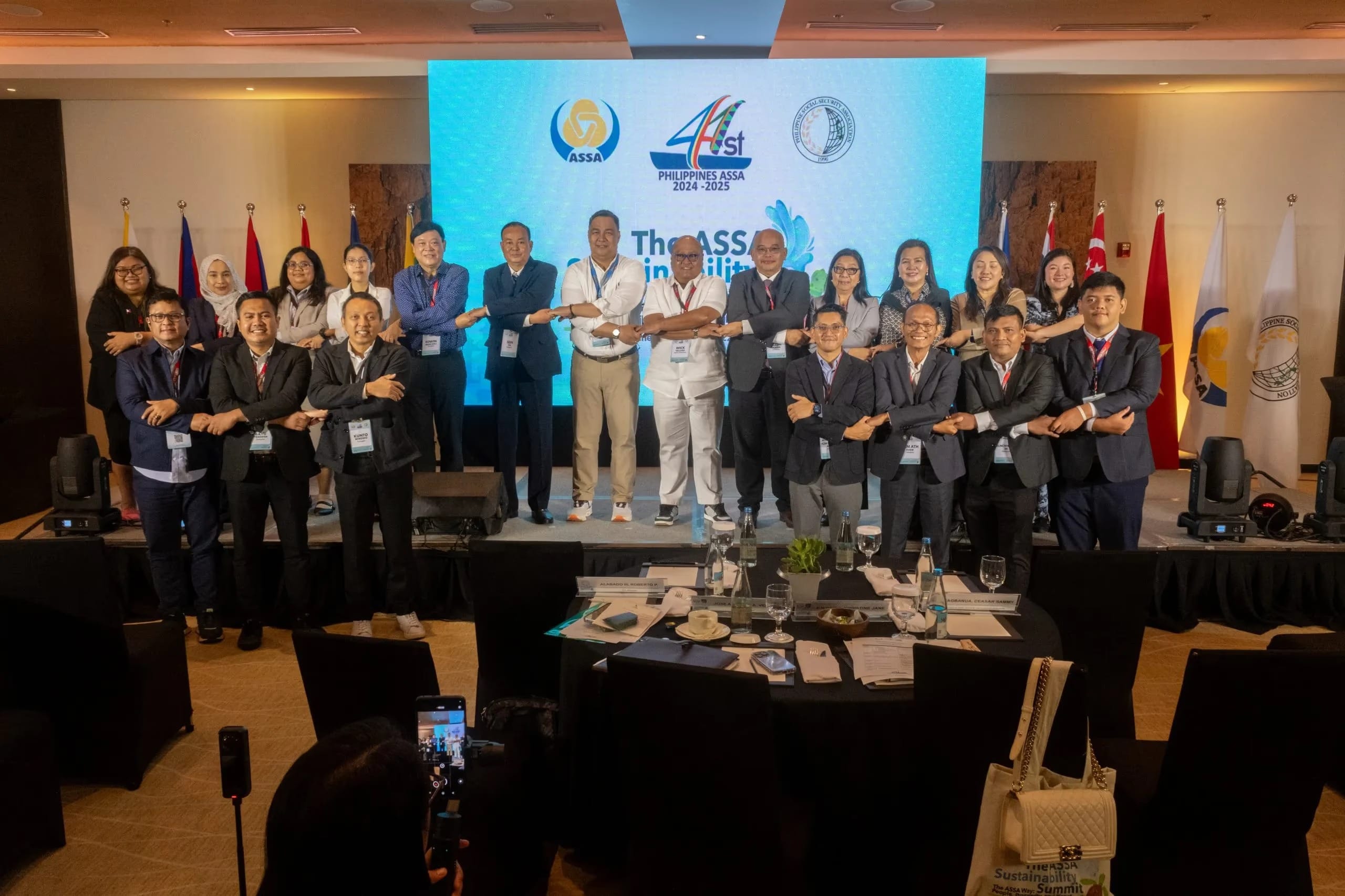 Group photo of delegates at The ASSA Sustainability Summit, held in Palawan, Philippines in March 2025 under the 41st Philippines ASSA 2024–2025 presidency. Approximately 20 participants pose on and in front of a stage, linking arms in the ASEAN way gesture. ASEAN member state flags are visible in the background alongside a large screen displaying the event branding.