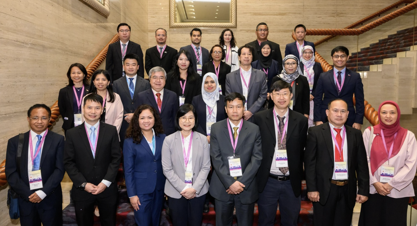 A group of people in formal attire, wearing lanyards, pose on a staircase with a mirrored wall in the background.