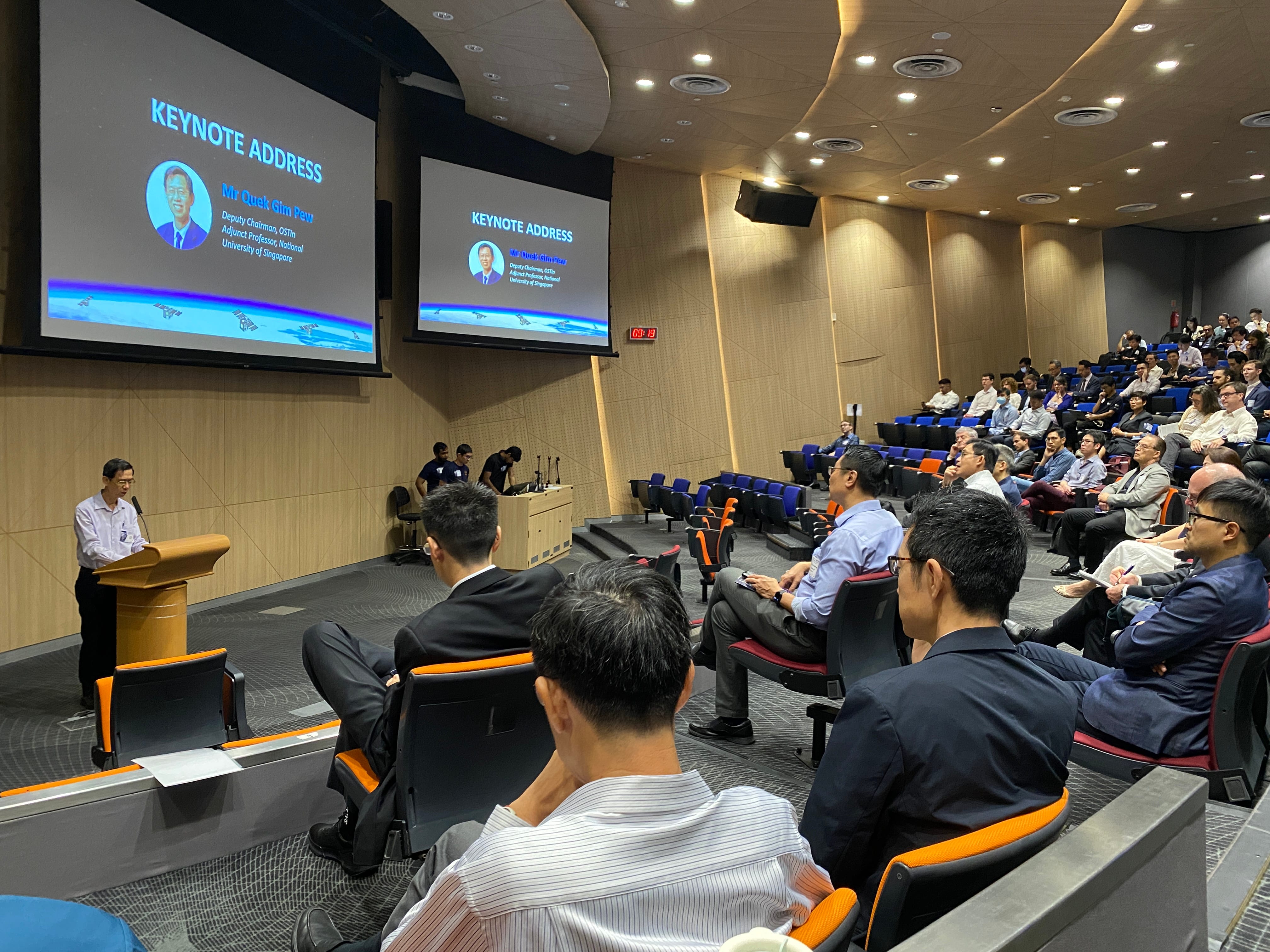 A speaker addresses an audience in an auditorium with a "Keynote Address" on two screens behind.