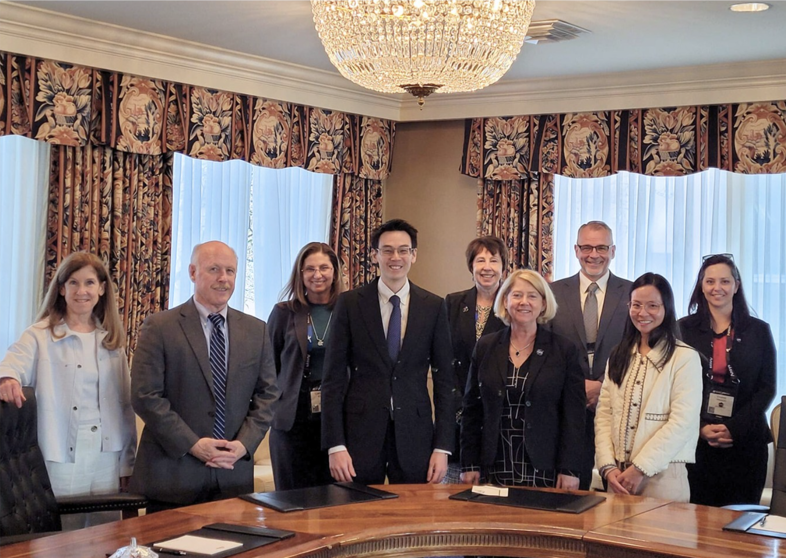 A group of people in formal attire standing around a large conference table under a chandelier in a room with patterned curtains.