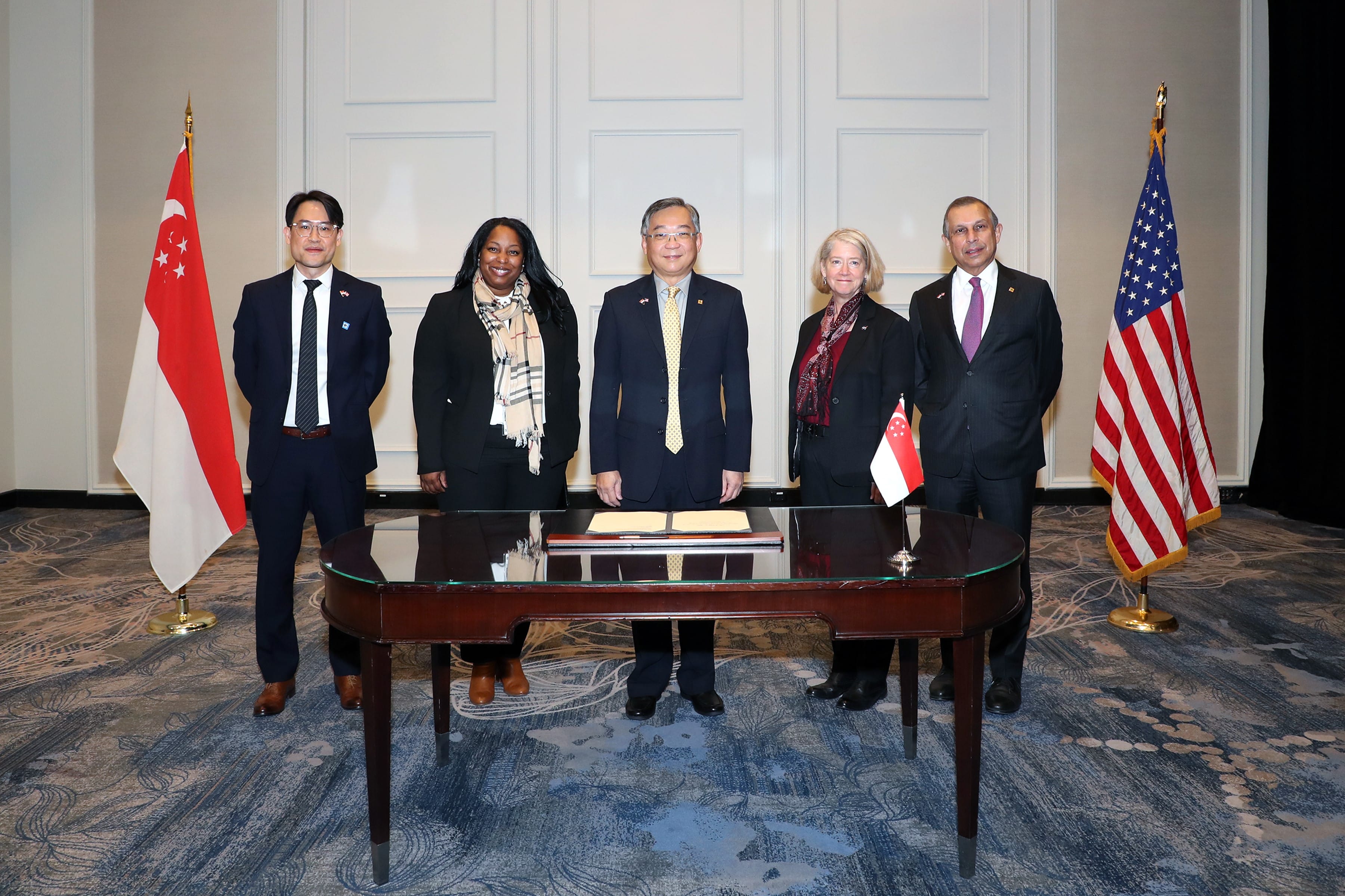 Five people stand behind a table with flags of Singapore and the USA flanking the group.