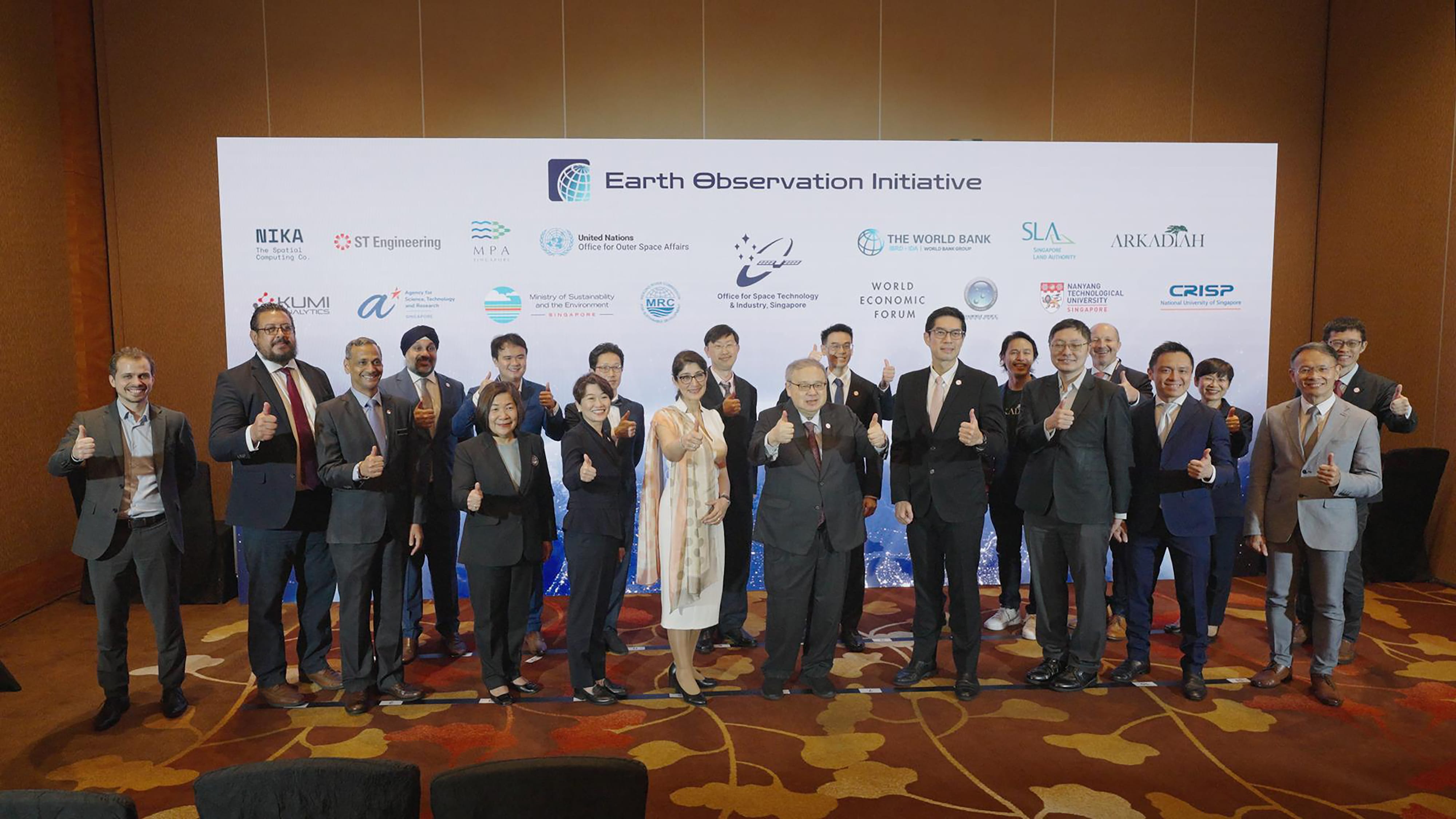 17 people in formal attire giving thumbs-up in front of a banner with logos and "Earth Observation Initiative" text.