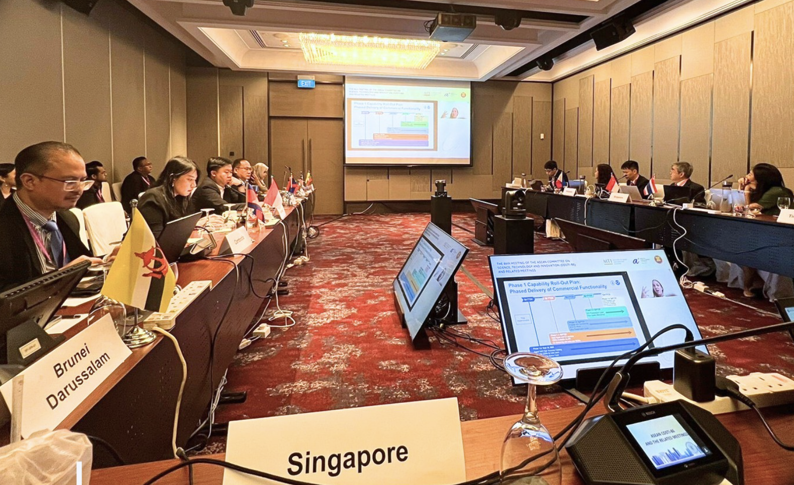 A conference room with delegates seated around tables with laptops and flags, viewing a presentation on multiple screens.