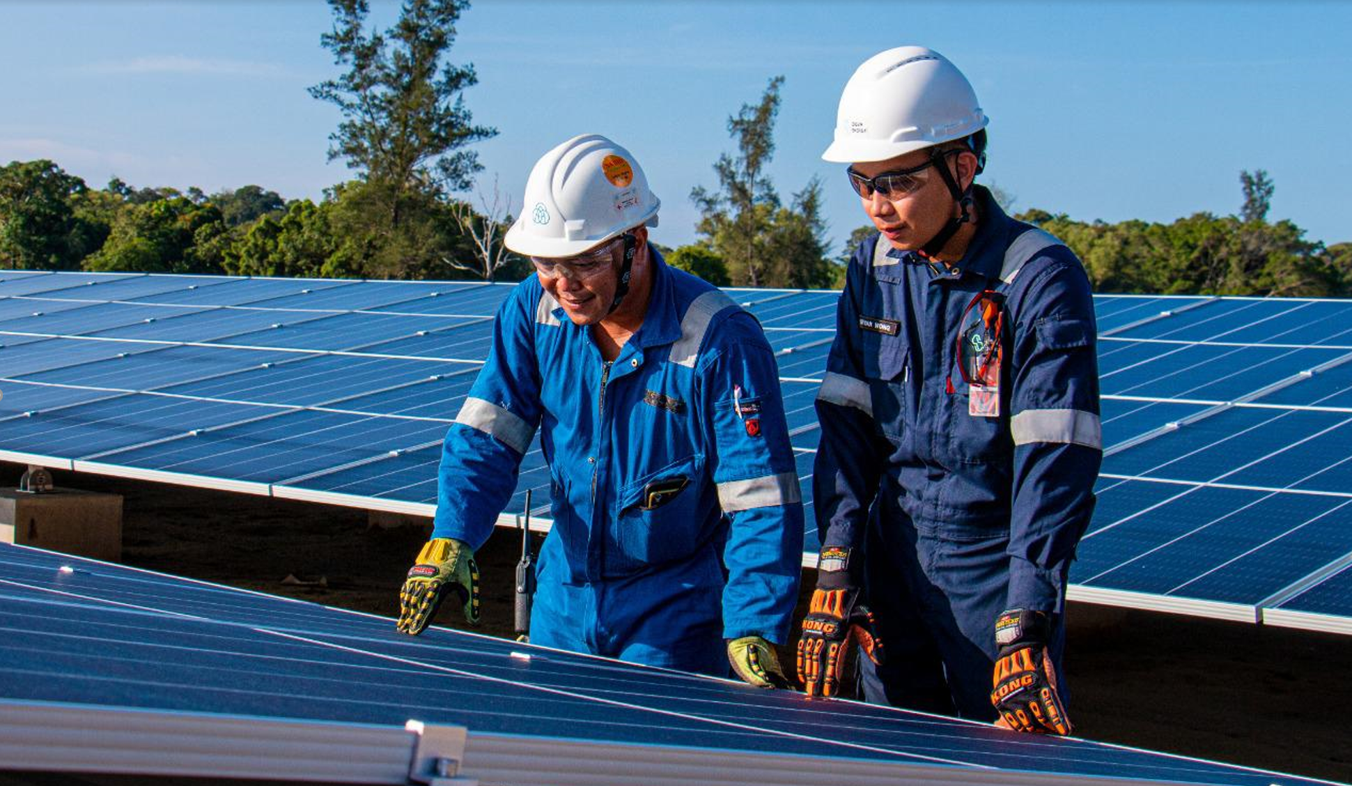 Two Eigen Energy workers in blue uniforms and white PPE helmets inspect solar panels in an outdoor setting.