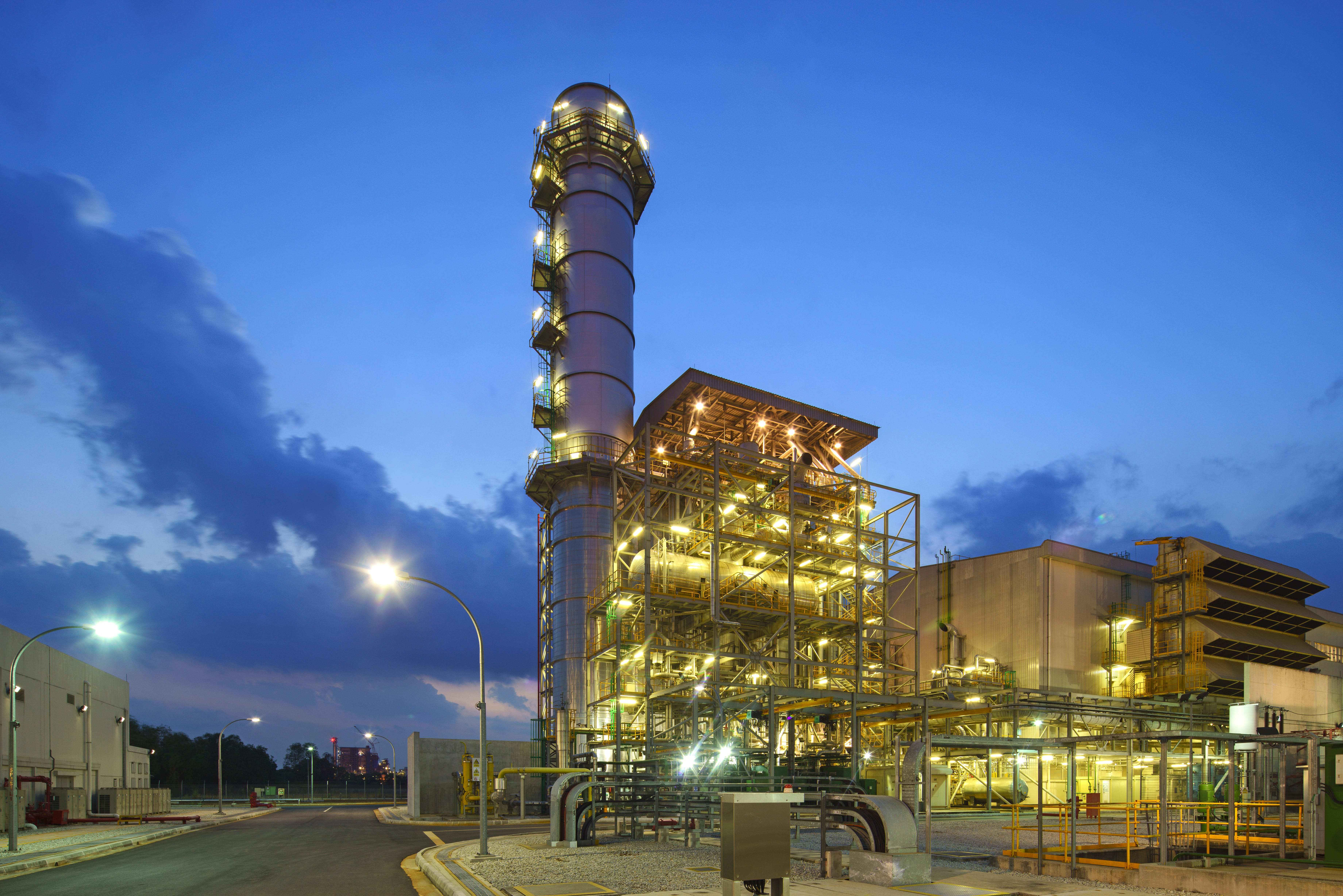 A power generation plant at dusk with bright lights highlighting a tower structure and metal framework against a blue sky.