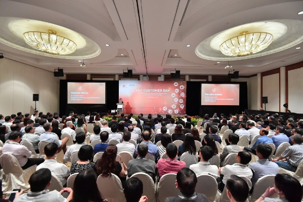 A large conference hall filled with attendees facing a stage with a speaker and red-themed screens reading "Customer Day."