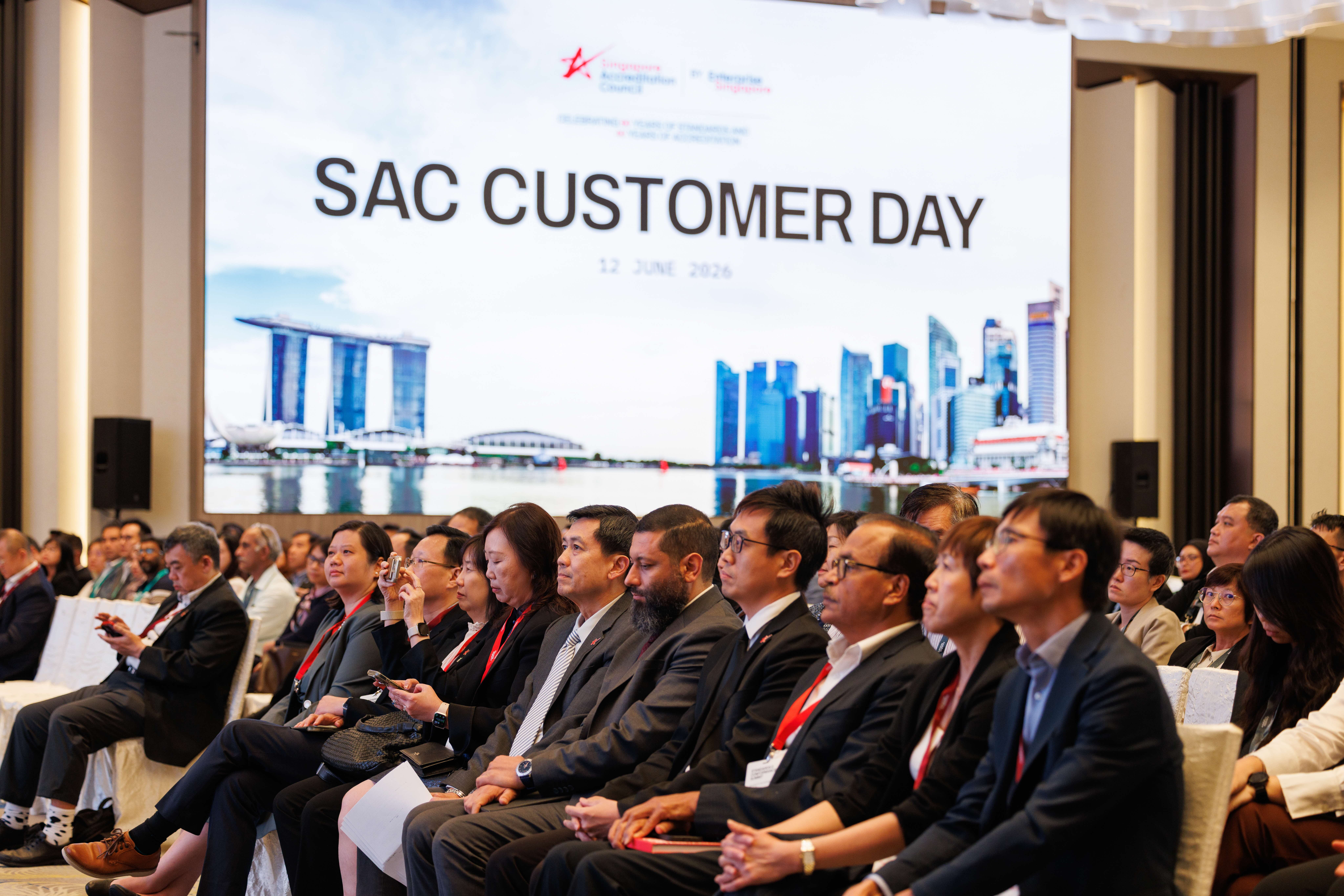 Audience seated at a conference facing a screen reading SAC CUSTOMER DAY with a city skyline backdrop.