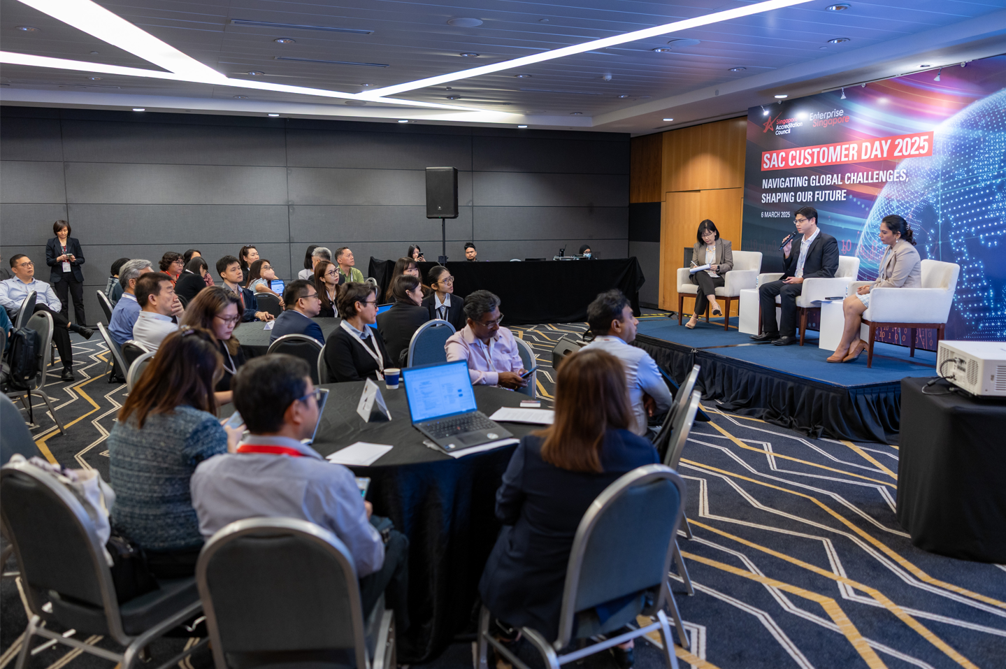 Three panelists sit on a stage at SAC Customer Day 2025 before a seated audience in a conference room.