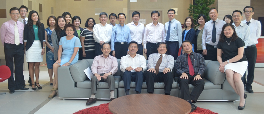 A large group of about 25 professionally dressed people posing together, with four seated on a grey sofa indoors.