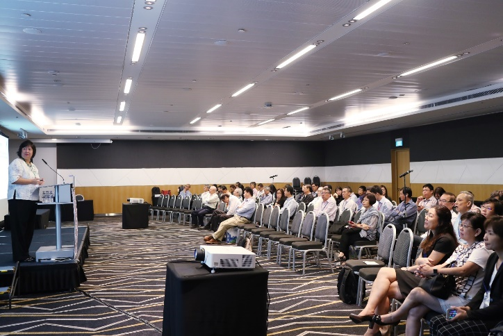 A woman presents at a podium to a large seated audience in a modern conference room.