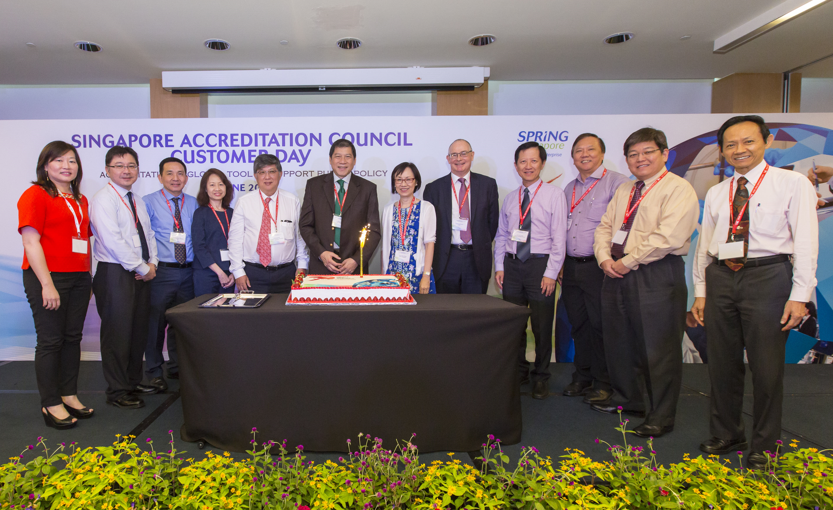 Eleven people pose behind a table with a large cake and sparkler at a Singapore Accreditation Council event.