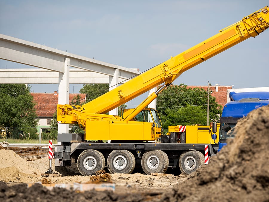 A large yellow mobile crane with an extended boom at a construction site near a concrete overpass.