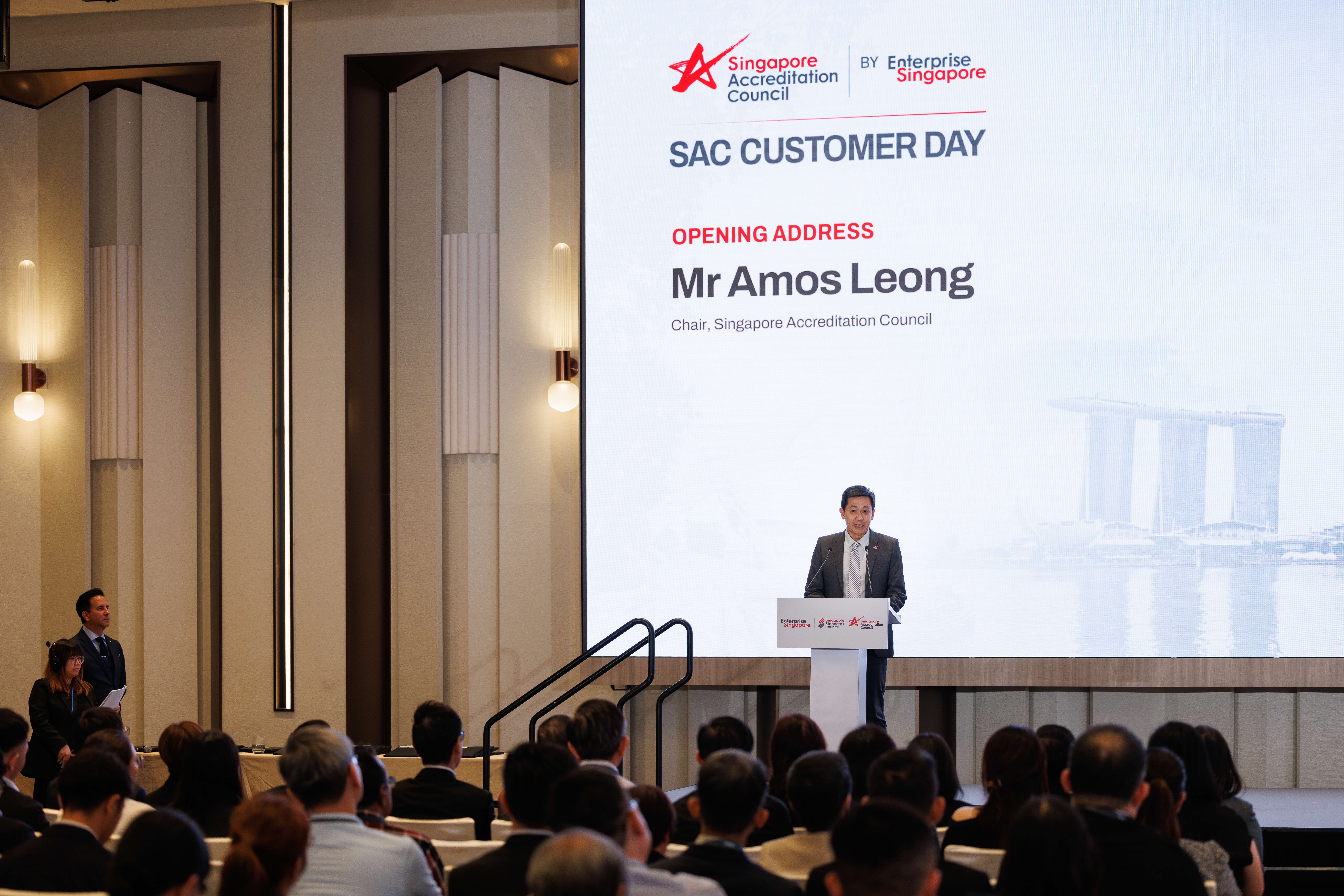 Man at podium speaks to seated audience; screen reads SAC Customer Day opening address Mr Amos Leong.