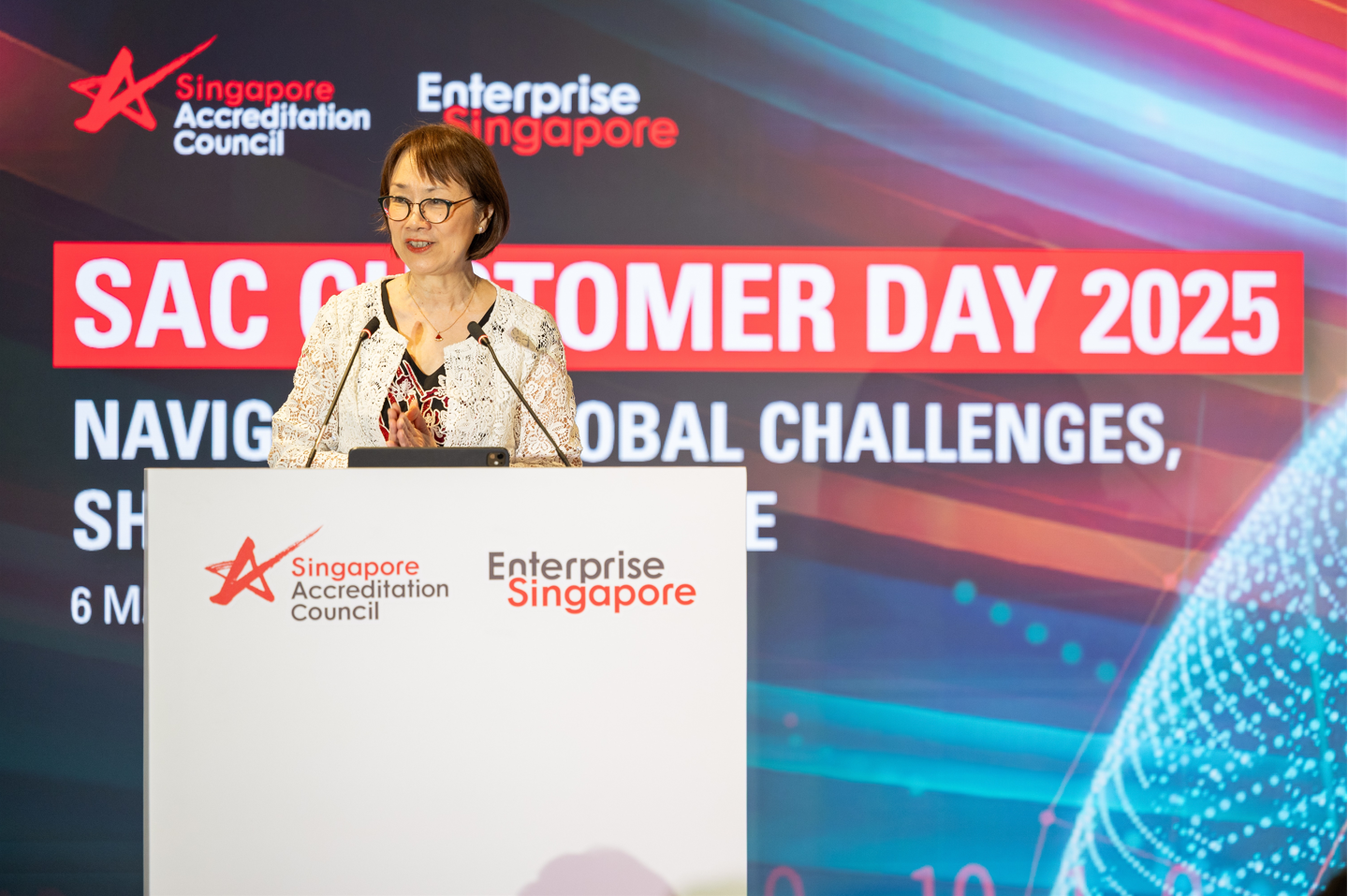 Woman in white lace jacket speaking at a podium at SAC Customer Day 2025, Enterprise Singapore event.