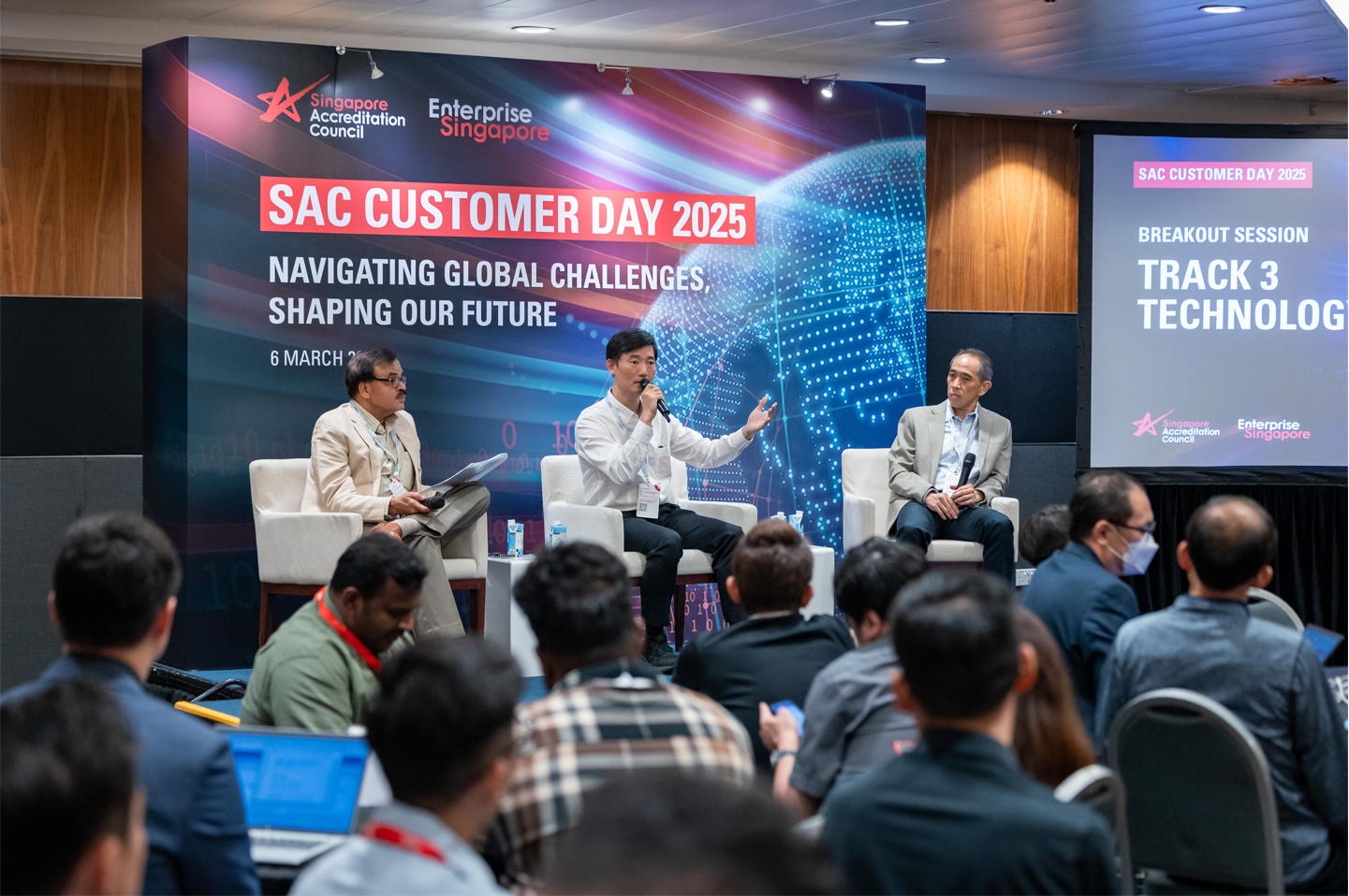 Three panelists seated on stage at SAC Customer Day 2025 conference, with audience members in the foreground.