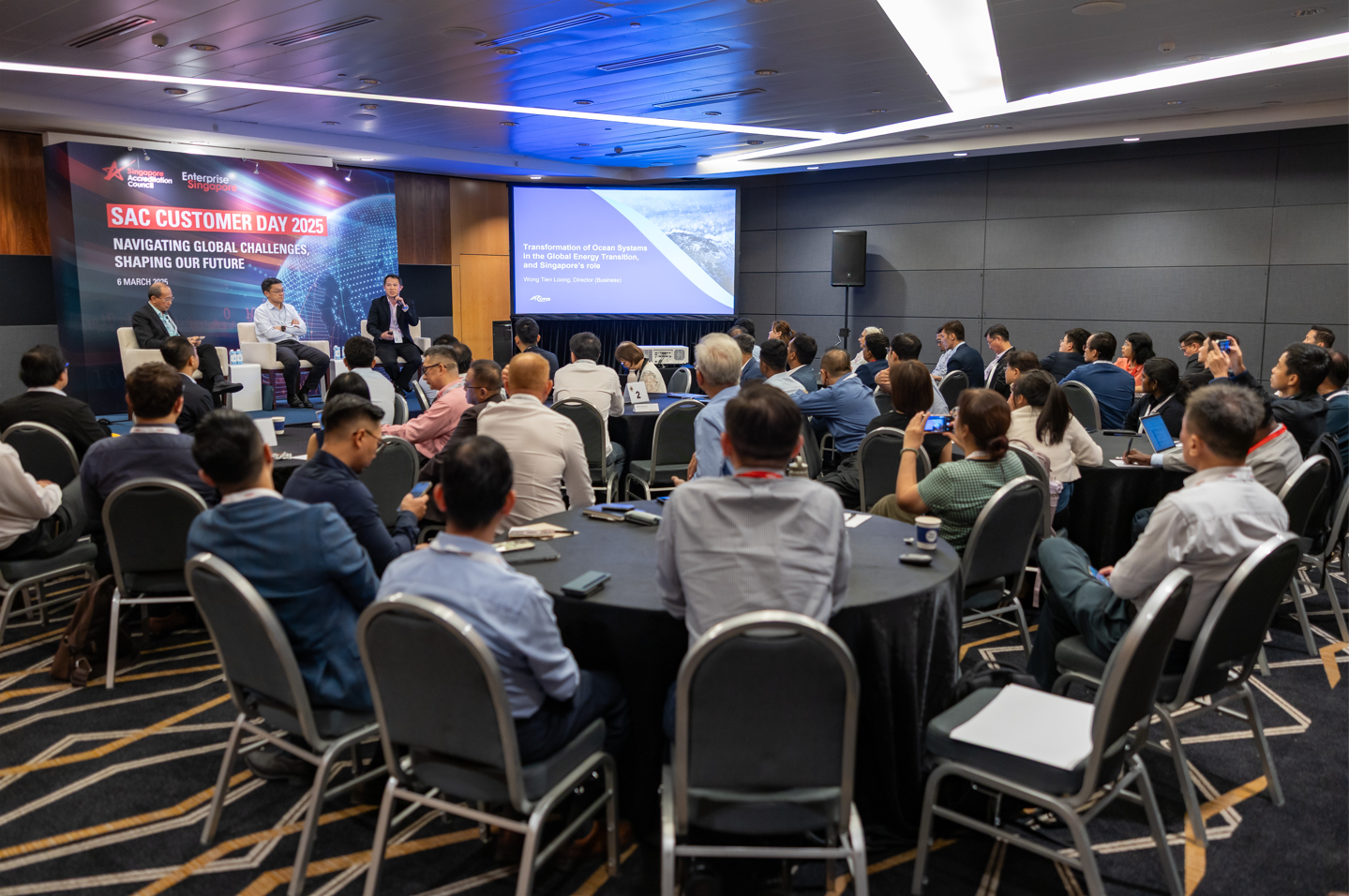 A conference room with attendees at round tables facing a panel and screen at SAC Customer Day 2025.