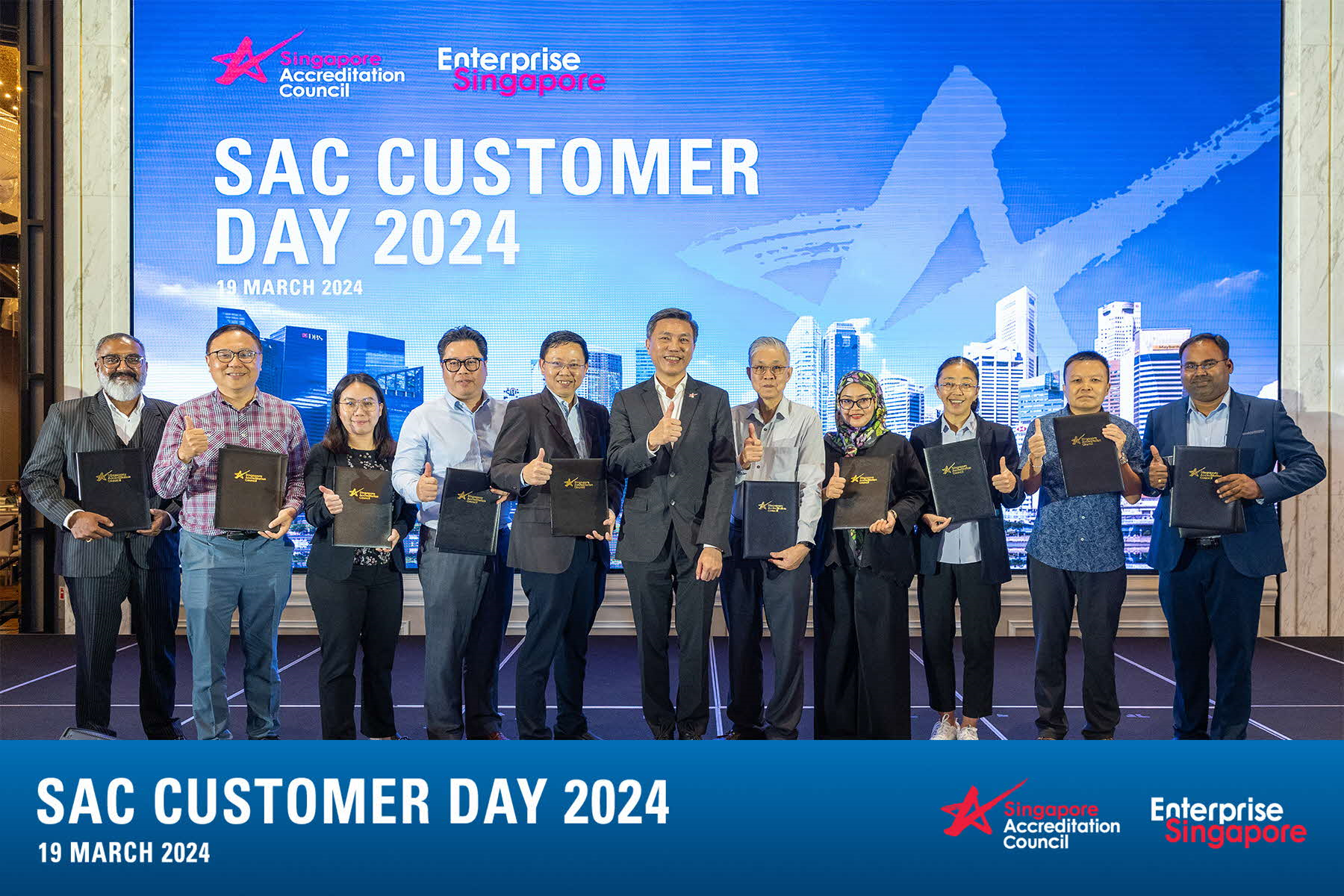 Eleven people stand holding black folders, giving thumbs up at SAC Customer Day 2024 on 19 March 2024.