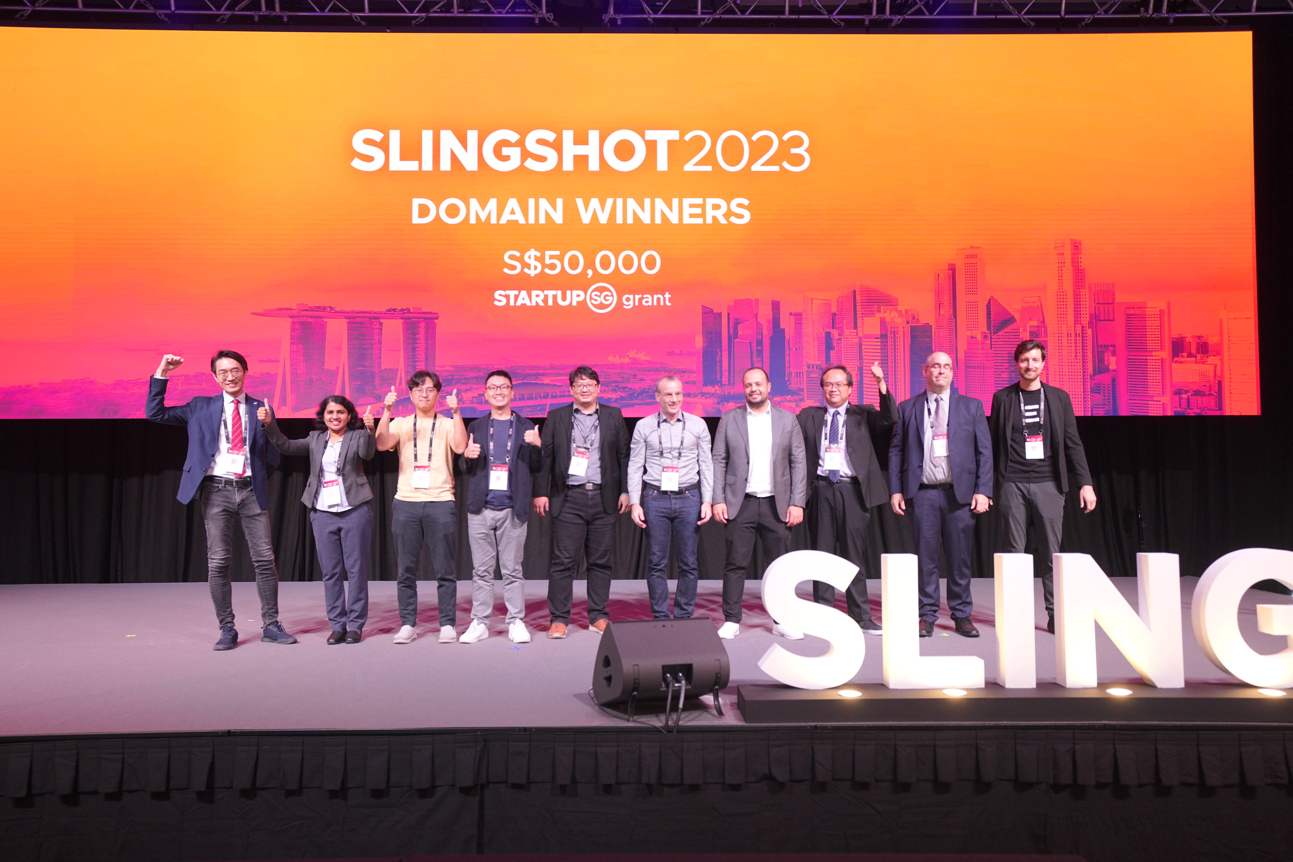 Eight people standing on a stage in front of a banner for SLINGSHOT 2023 Domain Winners, with an S$50,000 grant announcement.