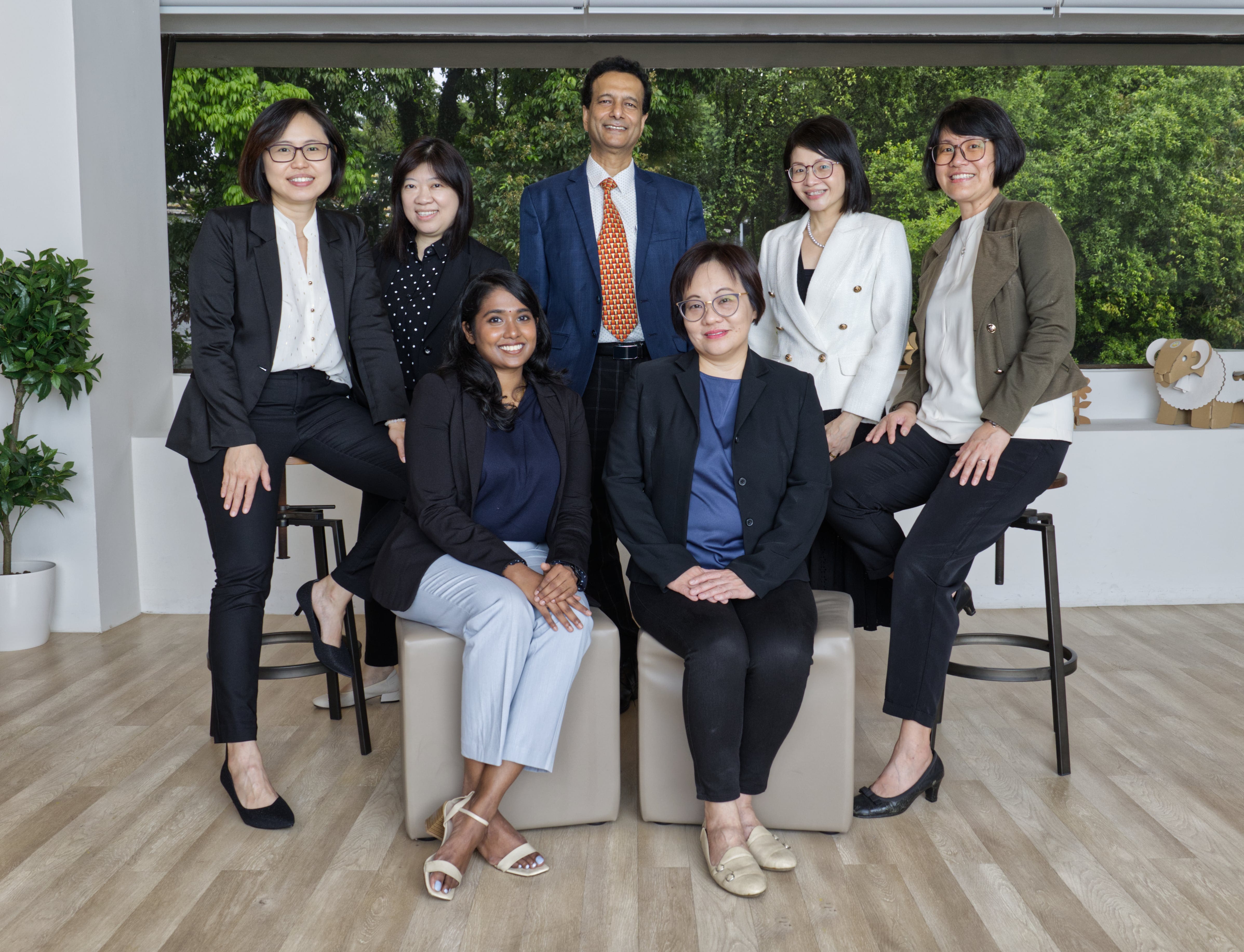 Seven people are posing indoors, with three standing and four sitting; some greenery is visible through the window in the background.