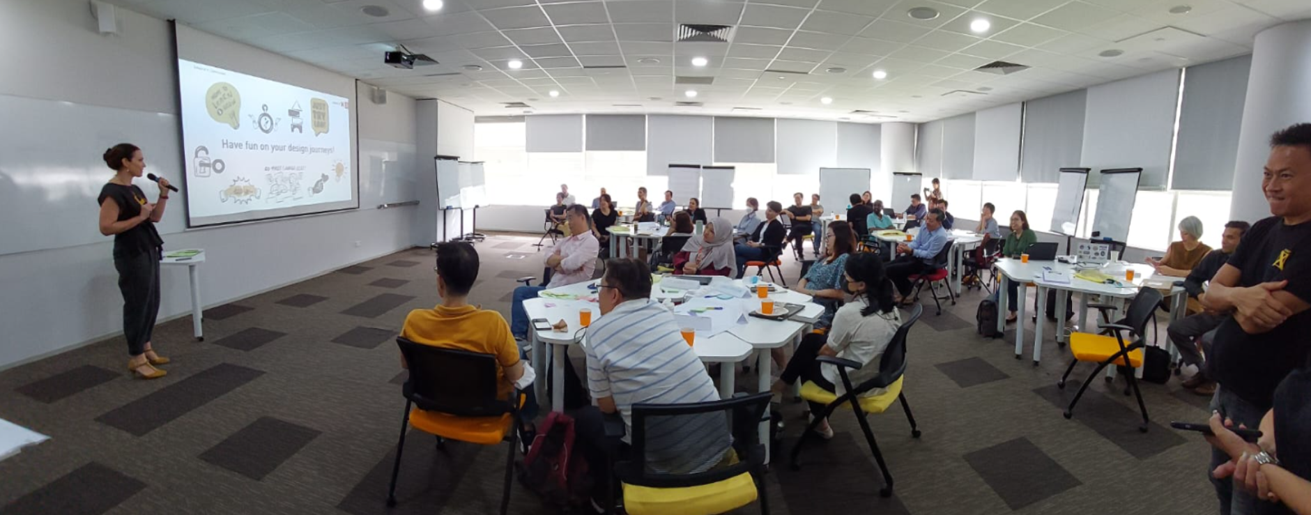 A wide-angle shot of a bright, modern classroom where a woman is presenting to a large group. She stands at the front with a microphone, gesturing toward a projection screen that reads 'Have fun on your design journeys!' Participants are seated at several white, modular tables arranged for group work, listening to the presentation.