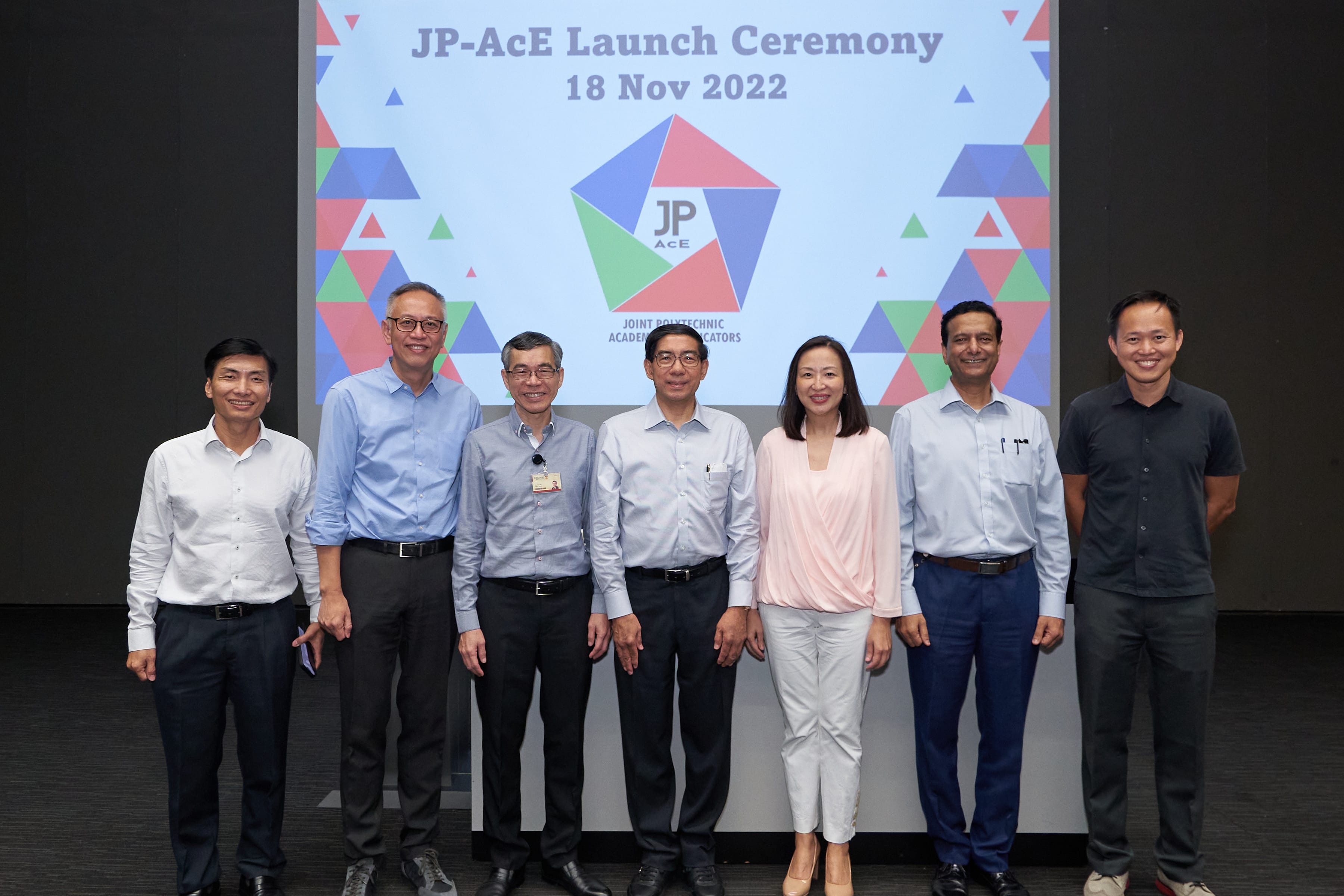 Seven smiling people stand in a line for a group photo. Behind them, a large screen displays the 'JP-AcE Launch Ceremony' title, the date '18 Nov 2022', and a colorful pentagon-shaped logo.