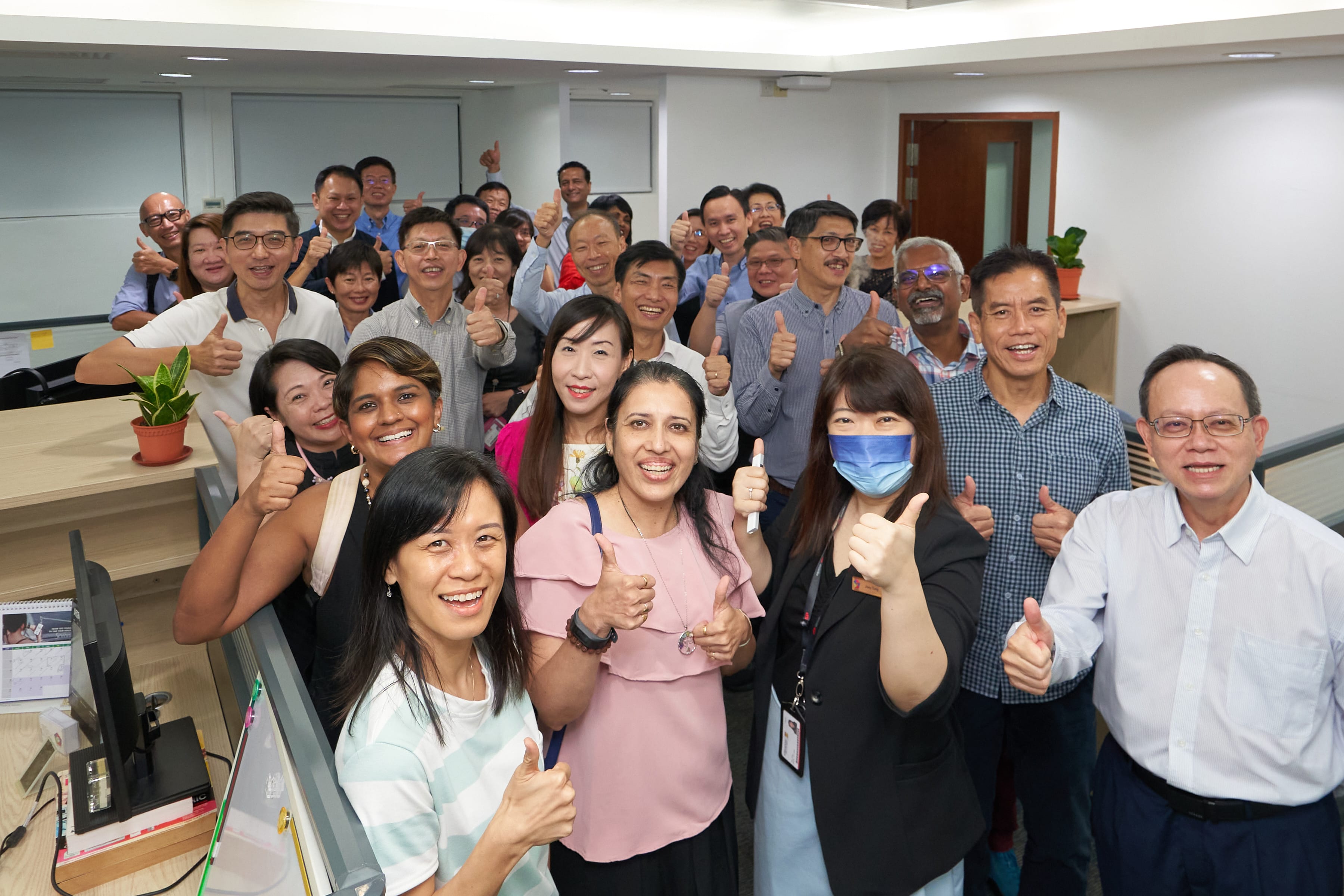 A large group of approximately 25 diverse people stand in rows, clustered within office cubicles. They are all smiling at the camera and most are giving thumbs-up gestures. One woman in the front wears a blue face mask